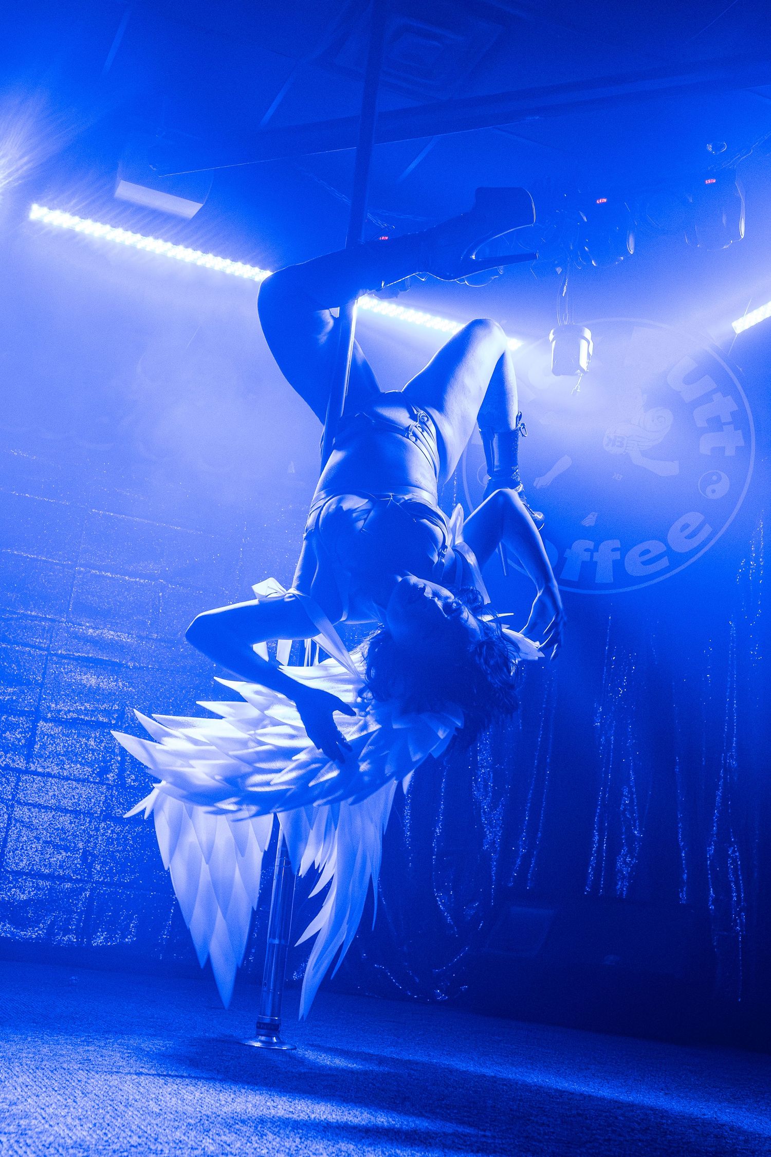 Acrobatic pole dancer performing an inverted move under dramatic blue stage lighting.