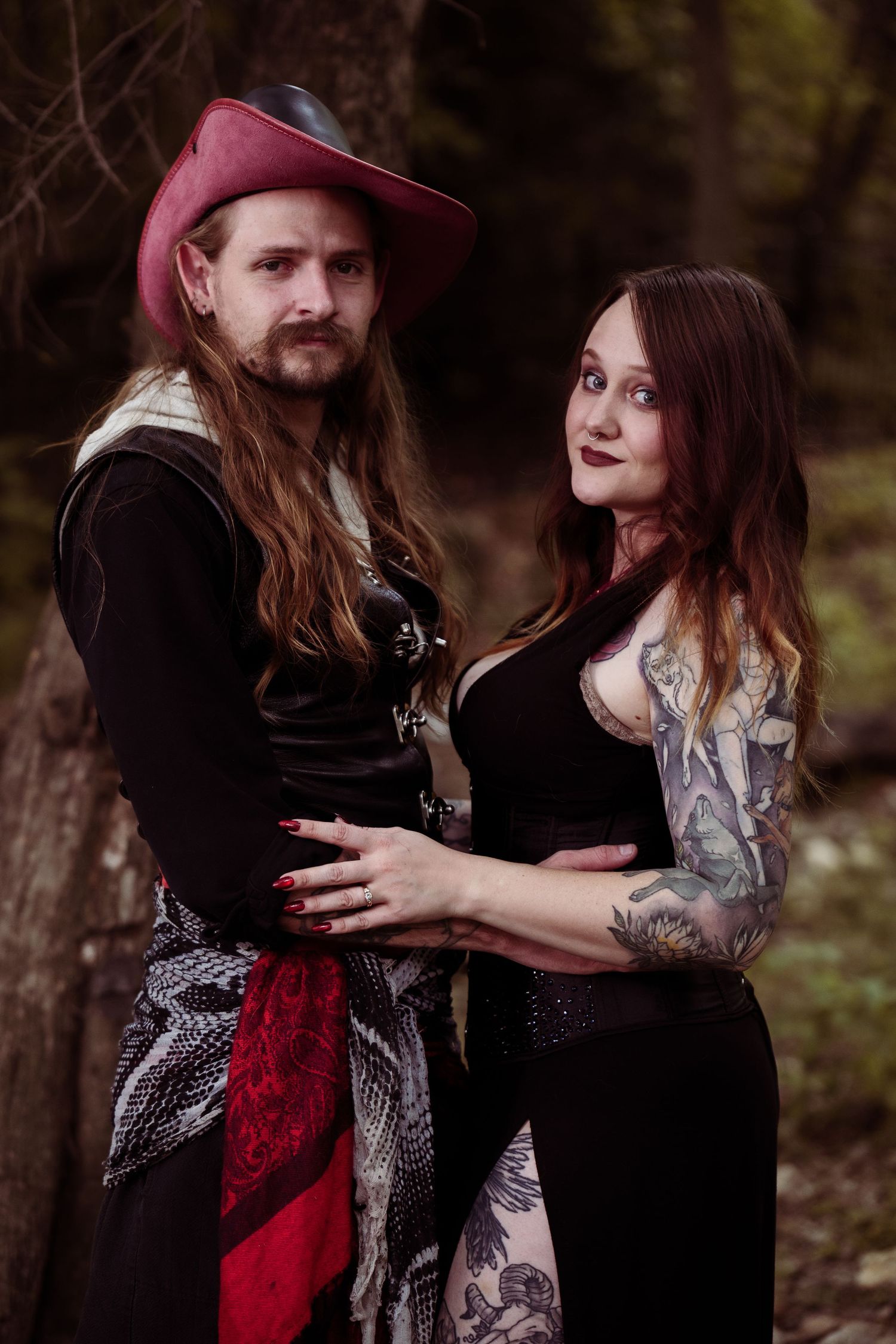 A couple in pirate costumes poses in a forest, the woman showing tattoos in a black dress, the man wearing a red hat.