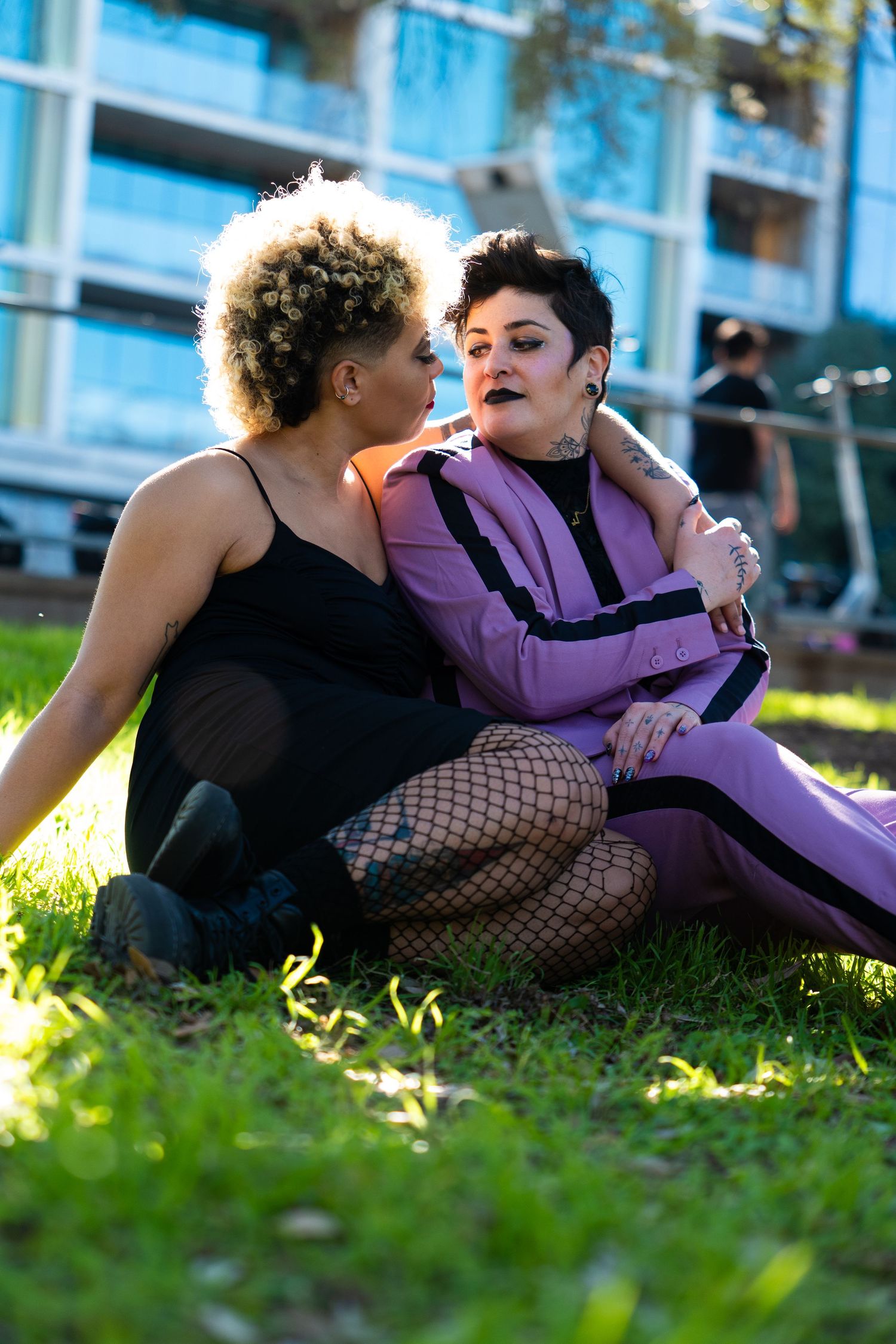 Two women sitting together on grass, one in black dress and one in purple suit, in urban park setting.