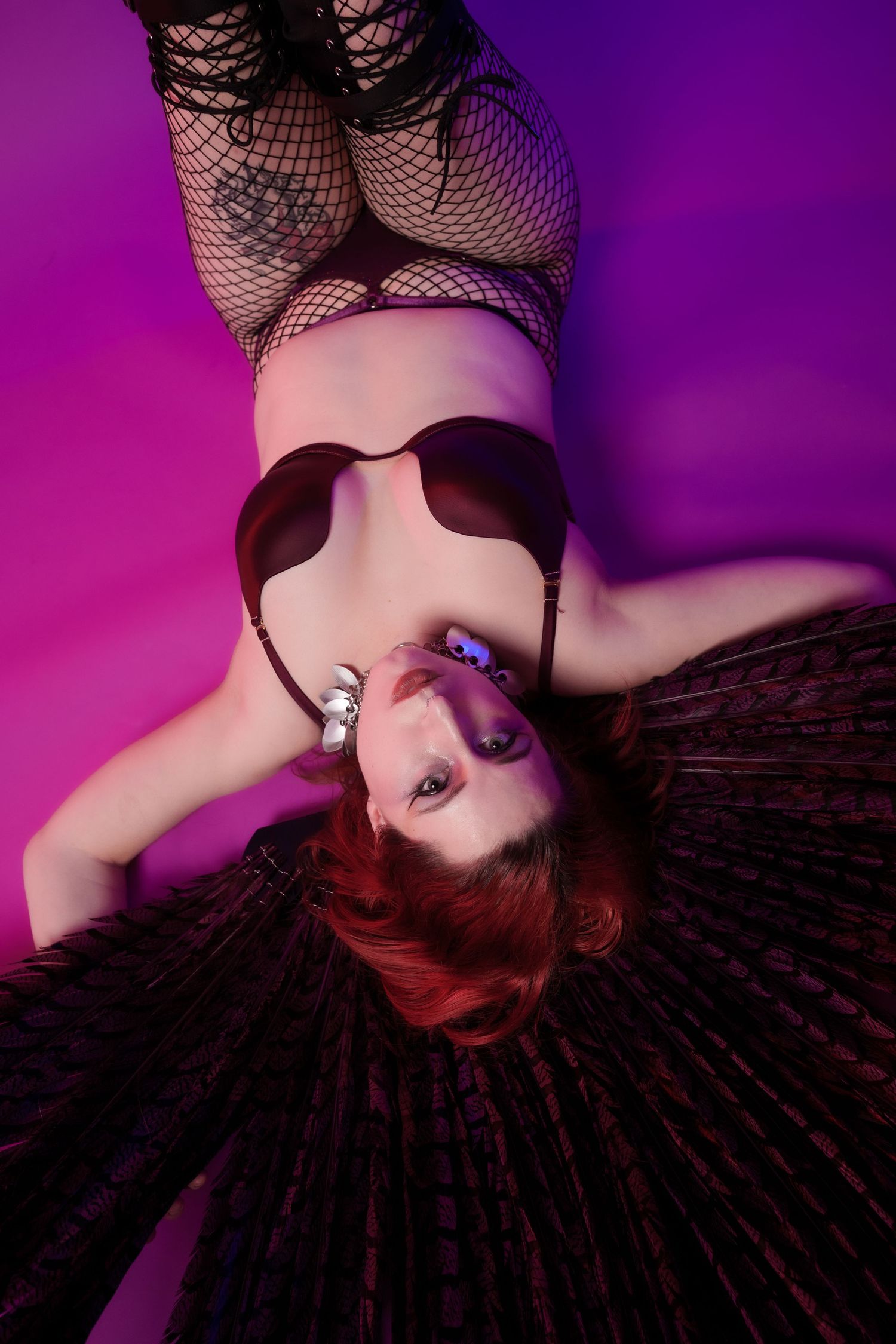 Woman in red hair and lingerie lying on purple background, wearing fishnet stockings and dark skirt.