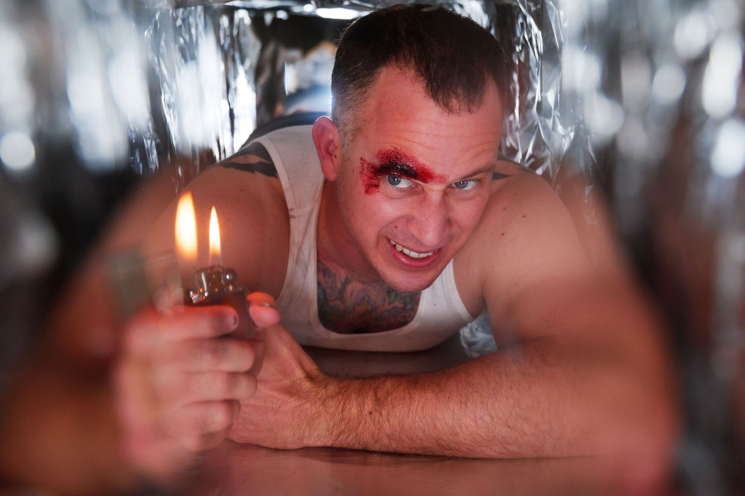 Man with fake blood wound holds a lighter while lying down surrounded by metallic tinsel.