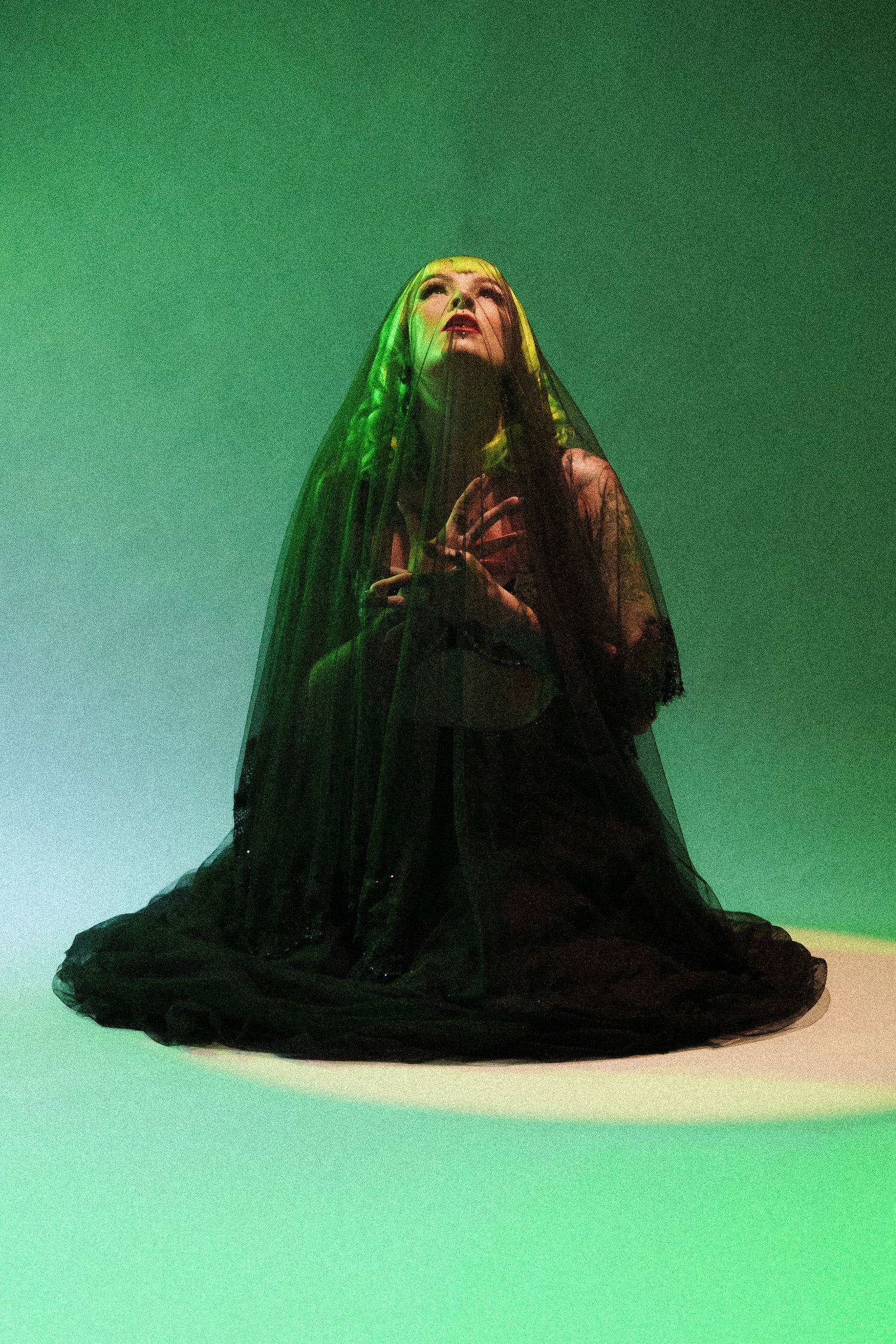 A person kneeling in a black veil and dark robes under dramatic green lighting in a studio setting.