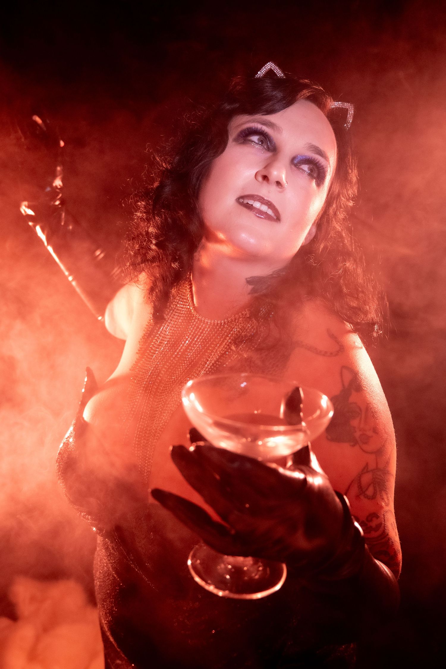 A tattooed woman in cat ears holds a cocktail glass while gazing upward in a red smoky dramatic scene.