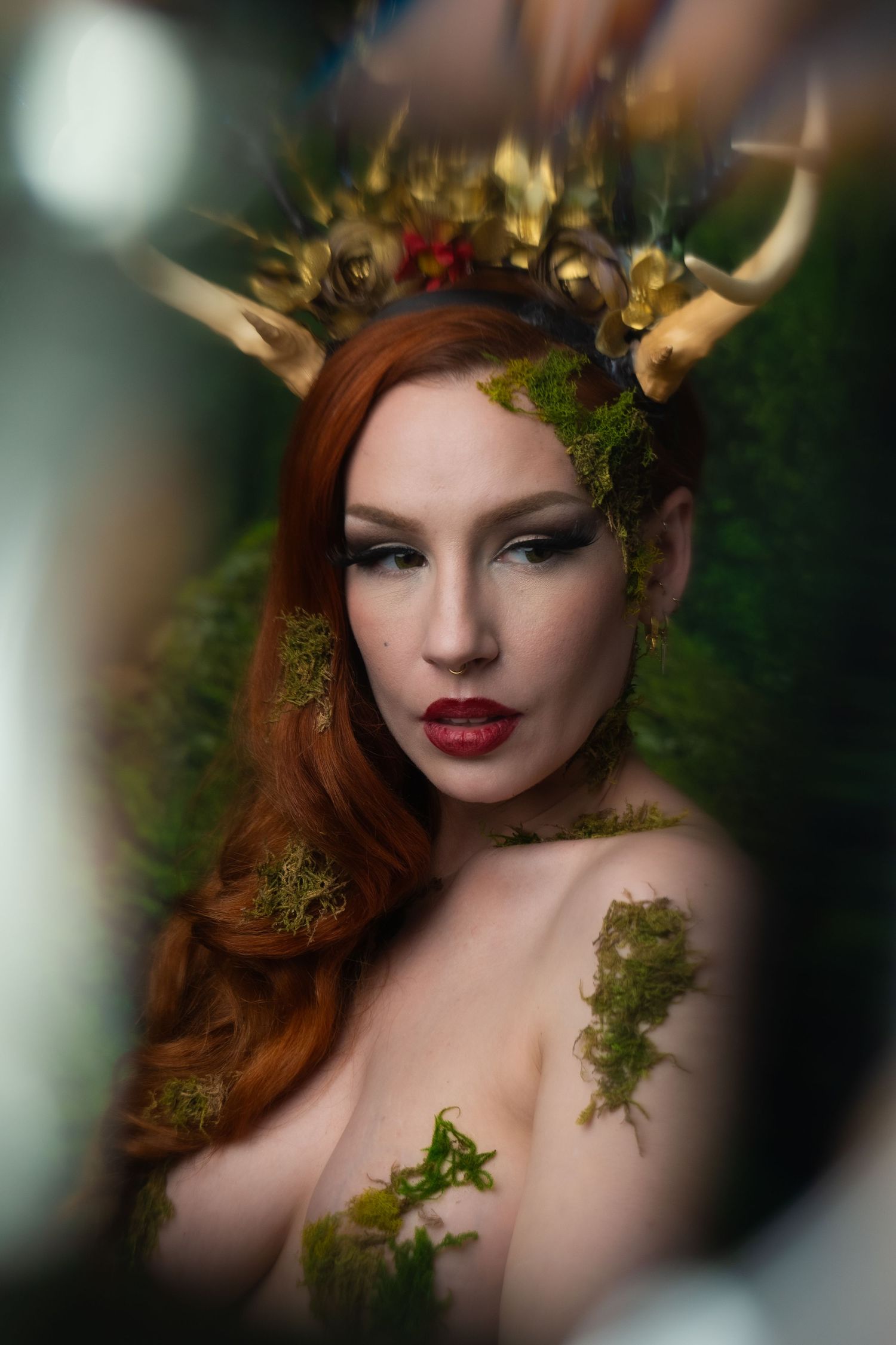 A redhead woman adorned with moss, antlers, and gold crown poses mystically in a nature-inspired fantasy portrait.
