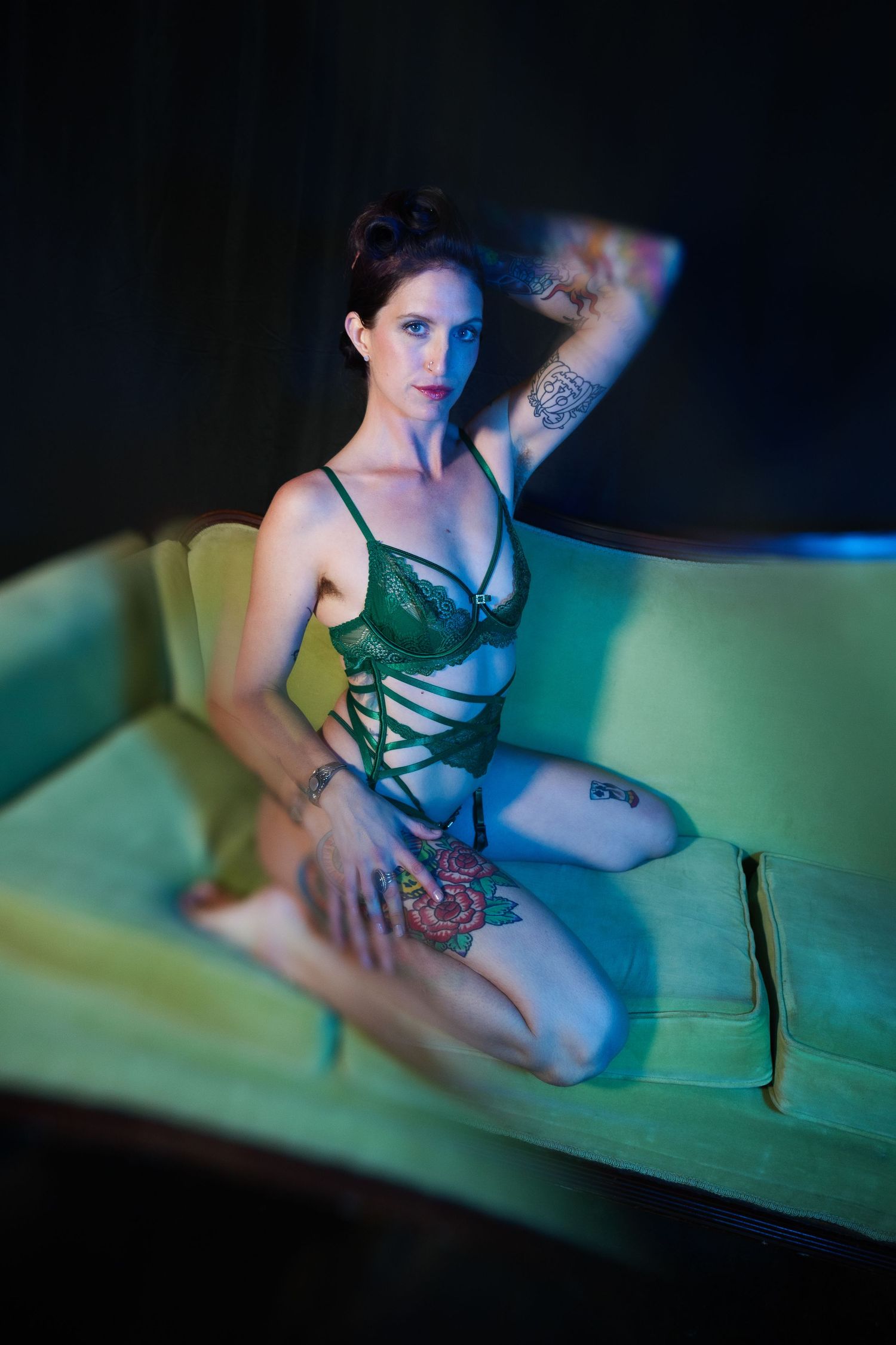 Tattooed woman in green lace lingerie kneeling on a vintage sofa under moody colored lighting.