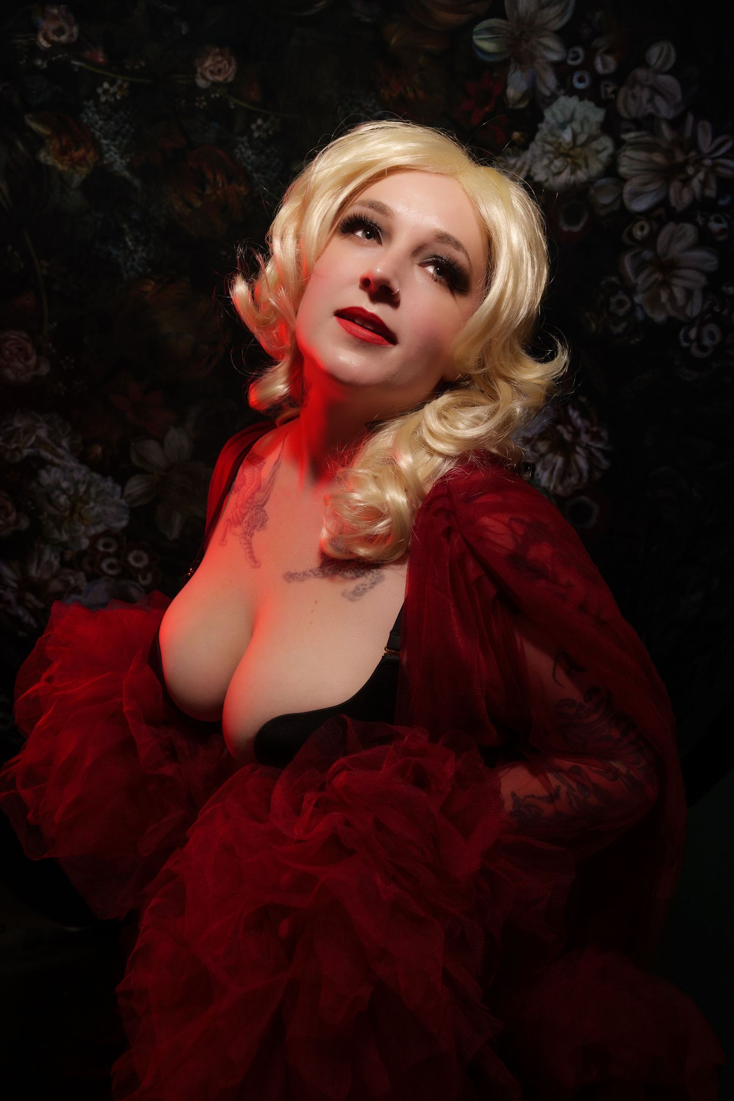 Blonde woman in red ruffled robe with red lips poses against dark floral background in dramatic lighting.