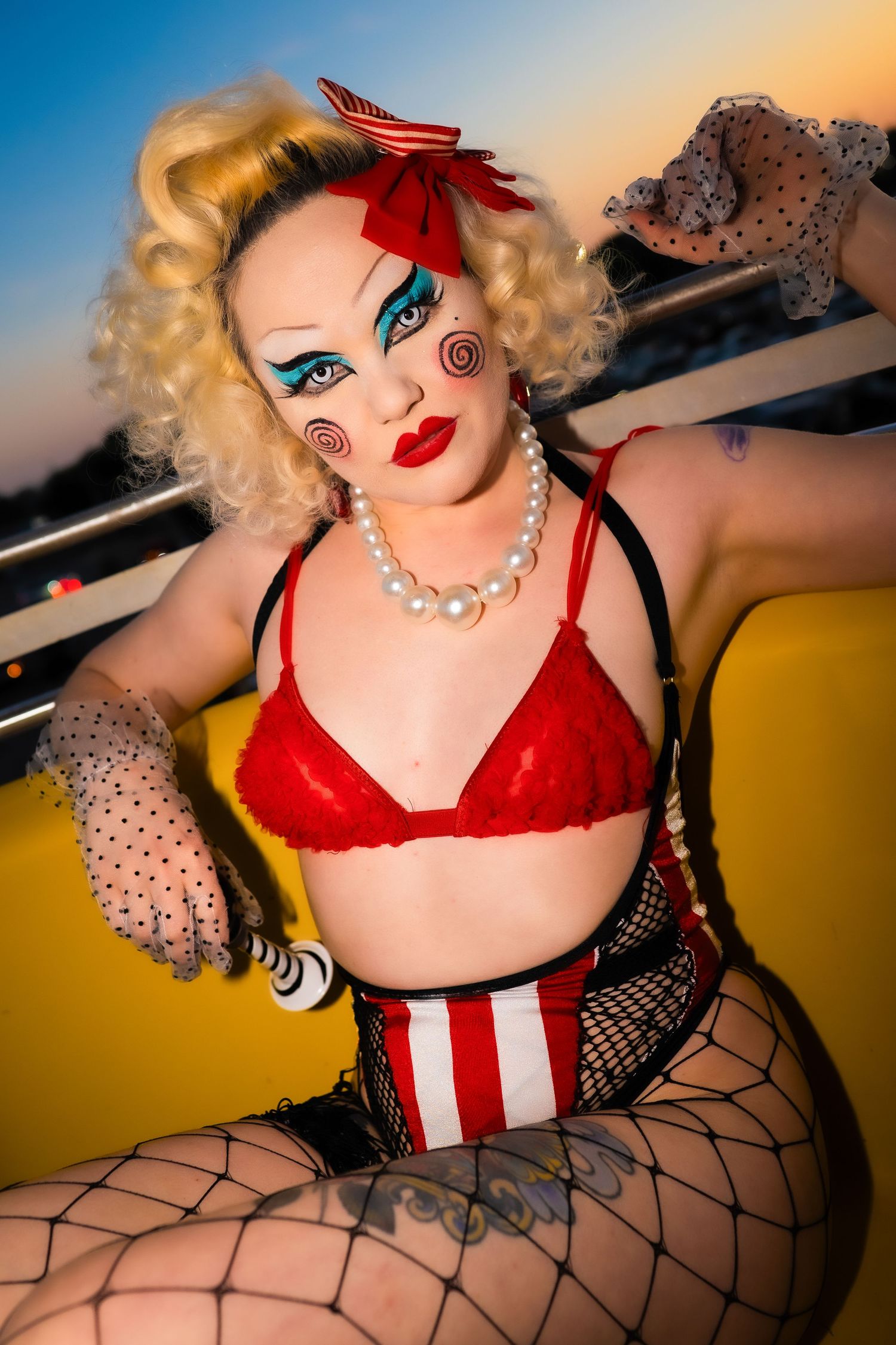 Woman in red bra with clown makeup and pearl necklace poses at sunset carnival.