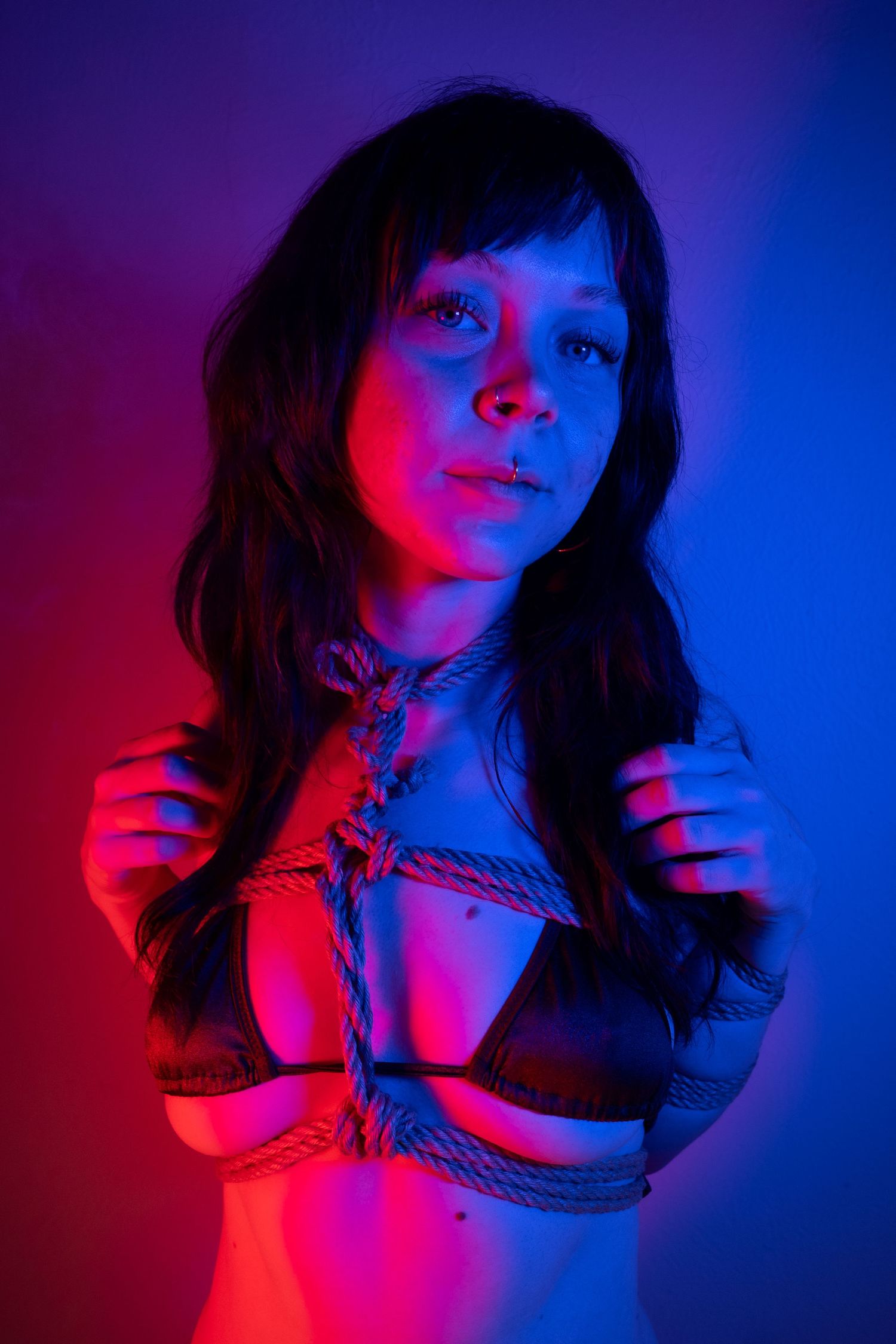 Young woman with rope bondage harness posing under dramatic red and blue studio lighting.