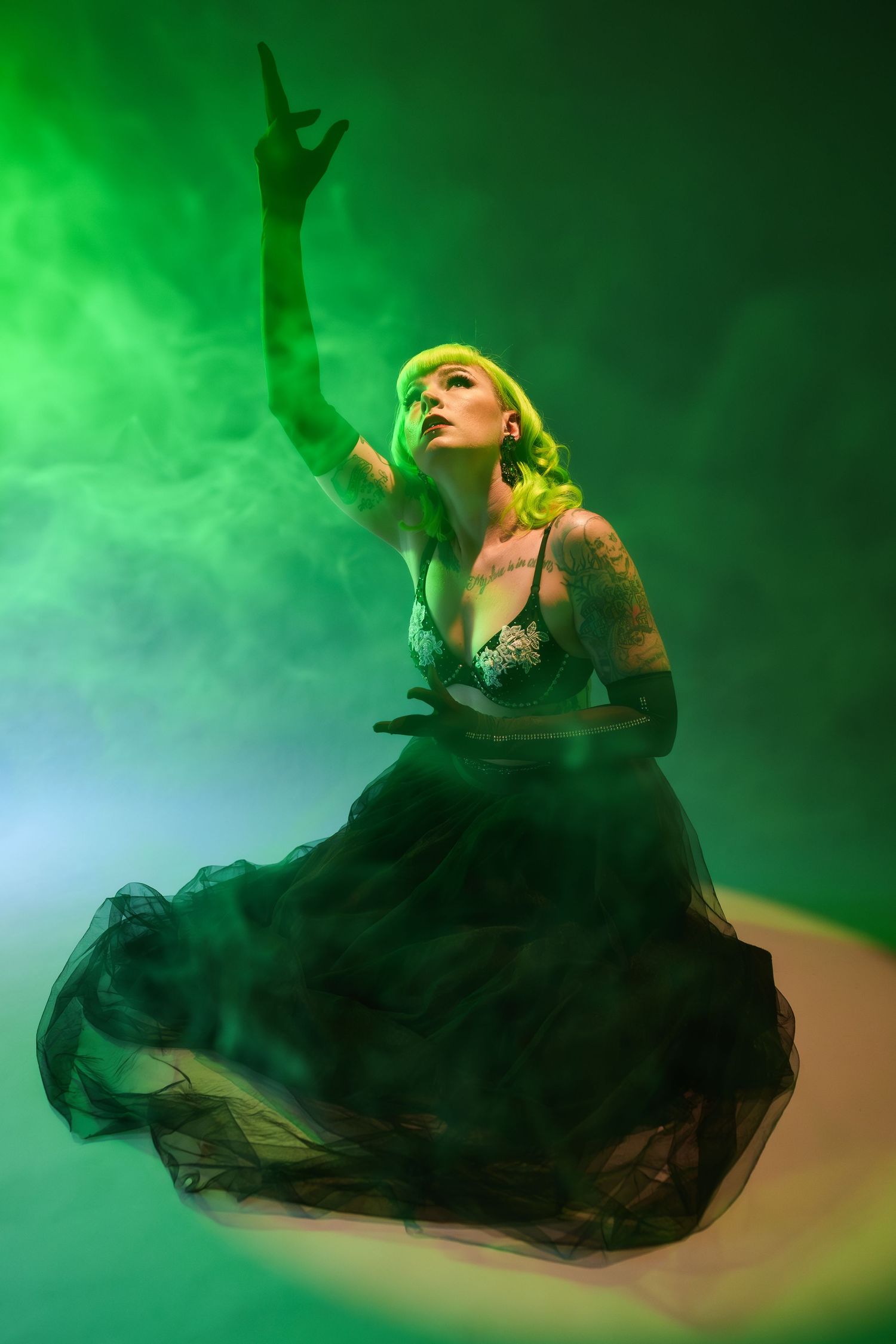 Tattooed blonde woman in black tulle gown poses dramatically in green smoky light with arm raised.