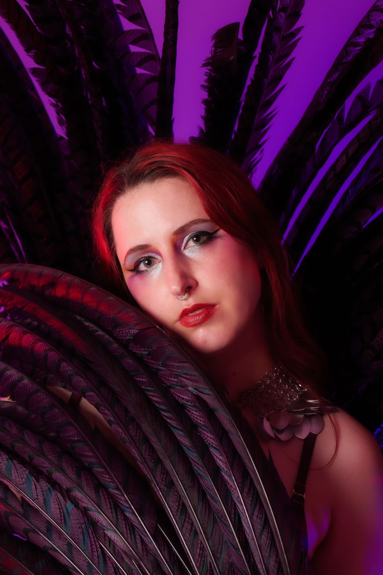 Young woman with red hair poses dramatically with large dark feathers against a purple background.