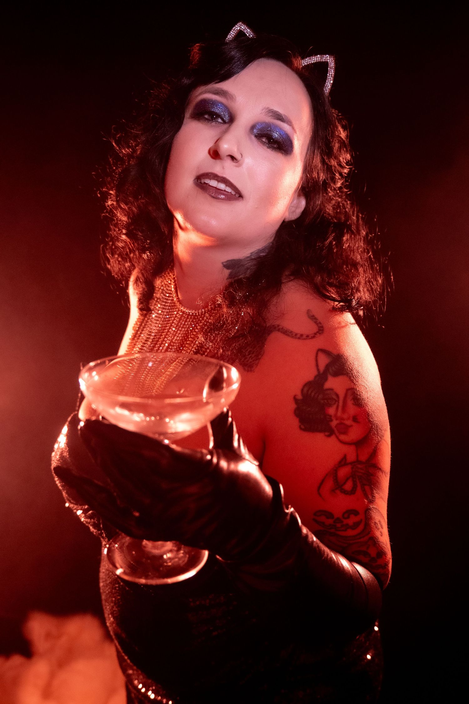 A curly-haired woman in cat ears holds a martini glass in dramatic red-lit smoky atmosphere with tattoos visible.