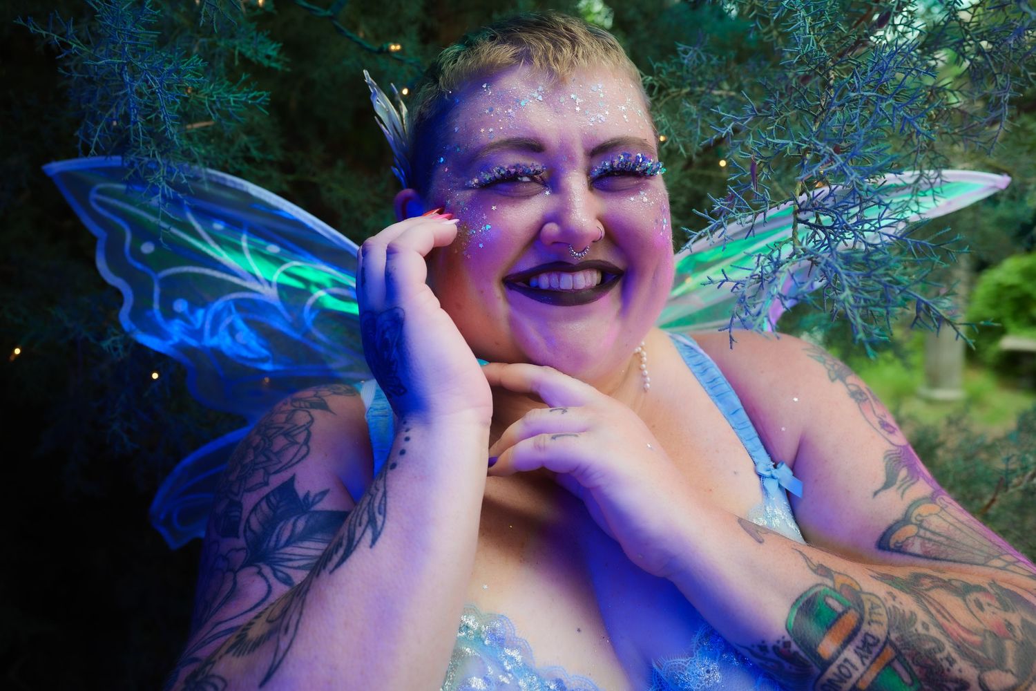 Smiling person in fairy costume with blue-green wings, fairy makeup, and tattoos posing outdoors in a magical forest setting.