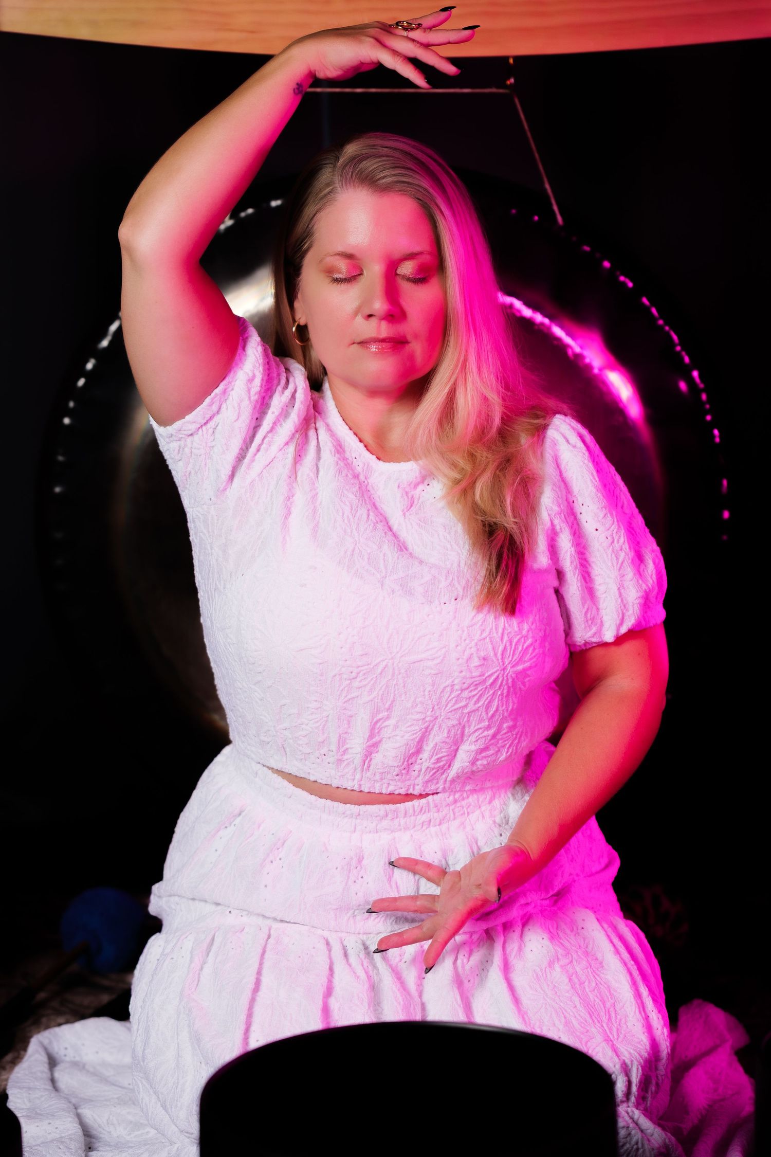Woman in white crop top and skirt poses gracefully under pink lighting against dark background.