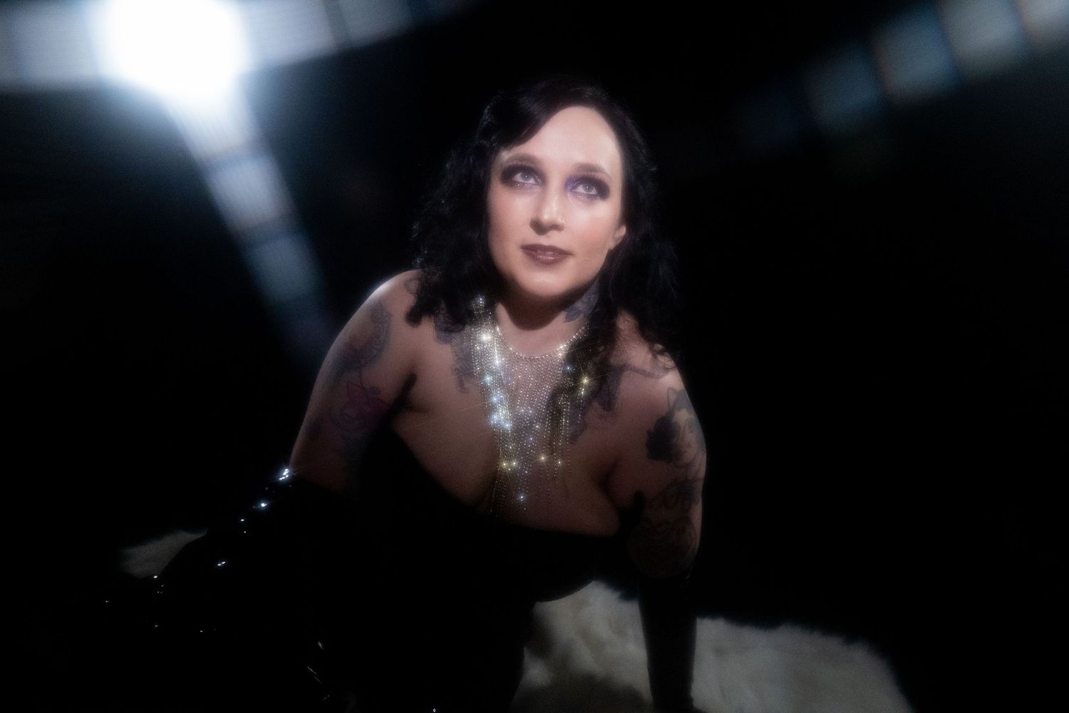 Woman with dark hair adorned in sparkling jewelry gazes upward in dramatic low-key lighting.