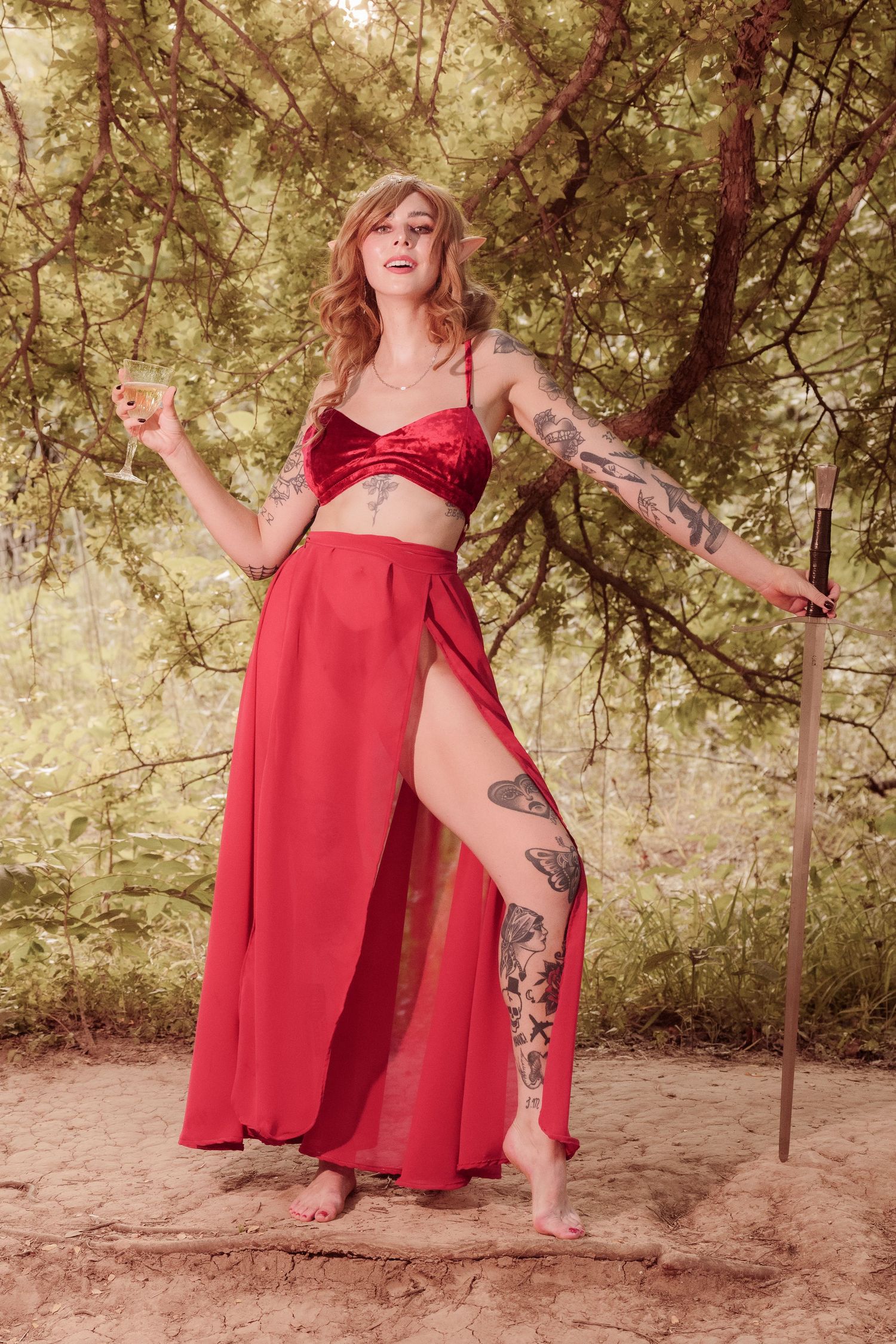 Woman in red velvet crop top and flowing skirt with tattoos poses outdoors holding a sword.