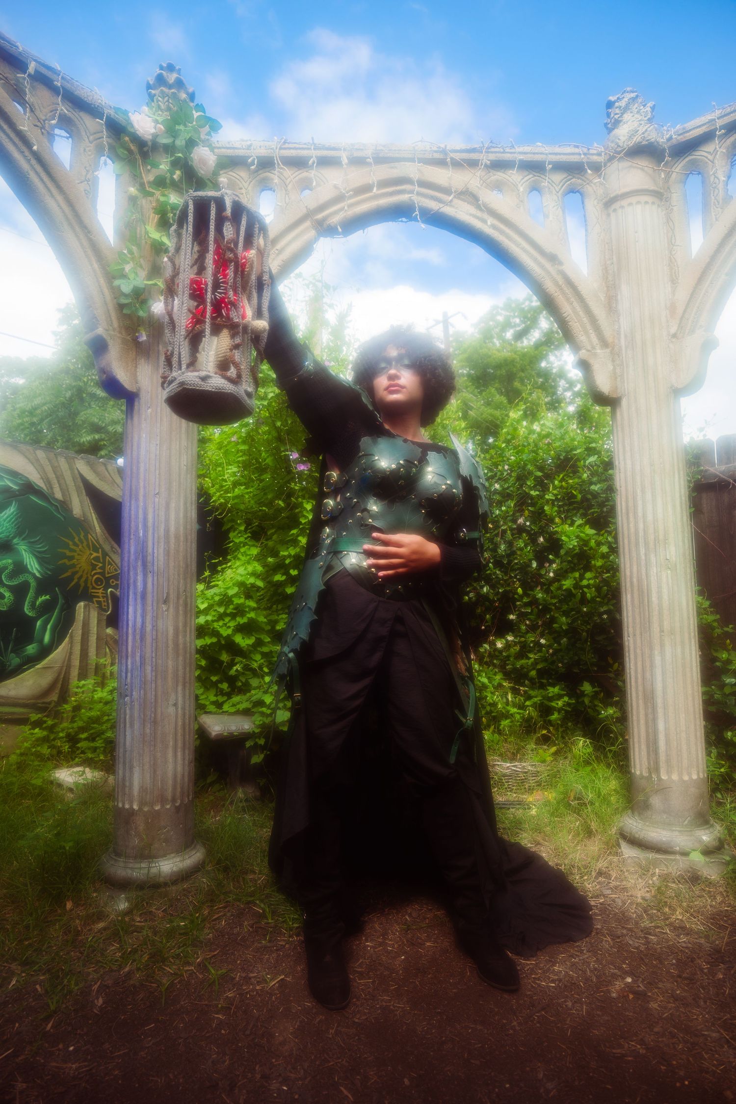 Woman in green corset and black skirt poses under gothic stone arch with decorative lantern in lush garden setting.