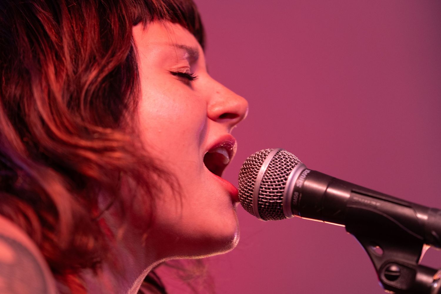 Young woman with eyes closed singing passionately into microphone bathed in warm pink and purple stage lighting.