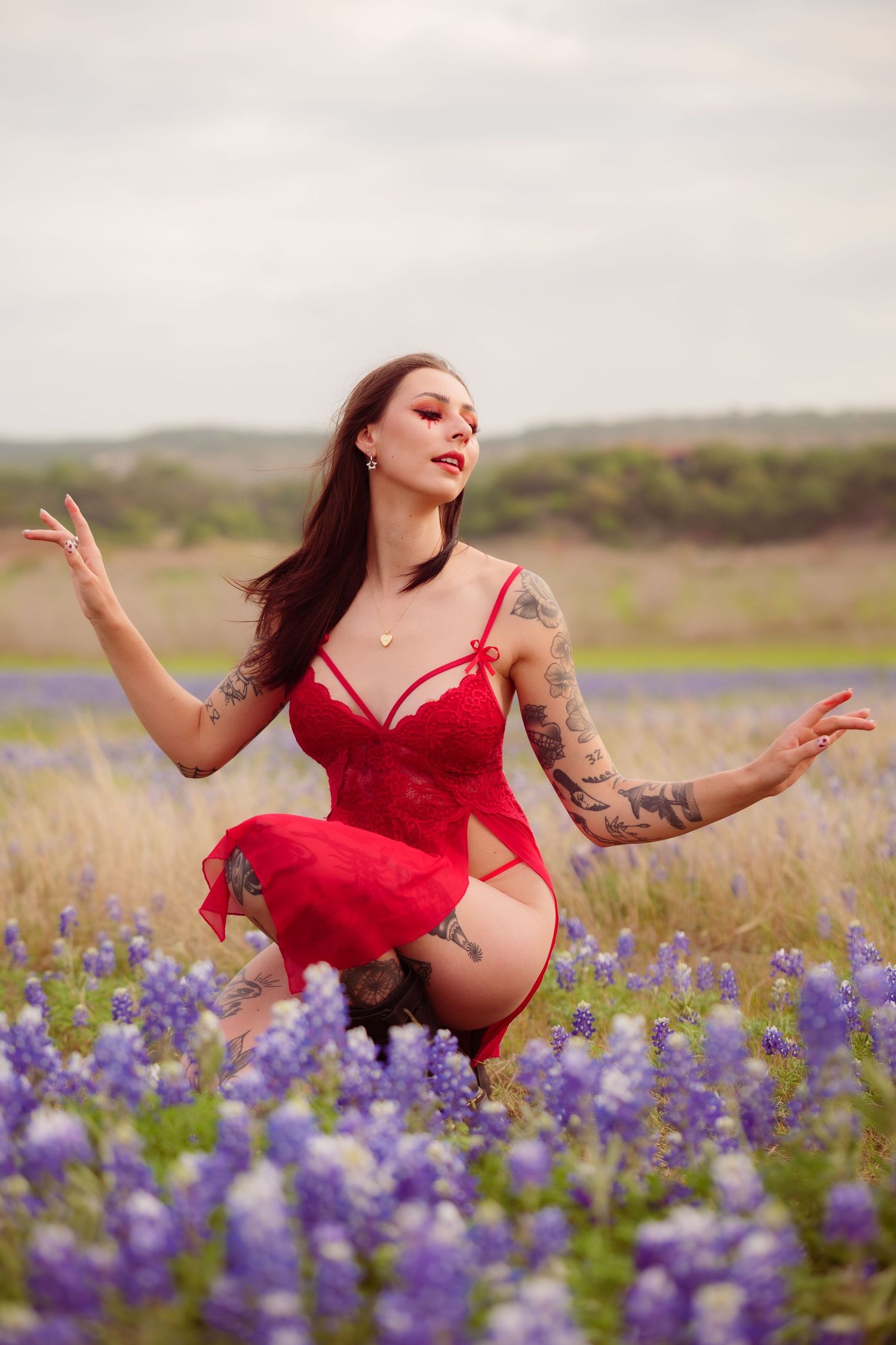 Tattooed brunette in red lace bodysuit poses gracefully among purple wildflowers outdoors.