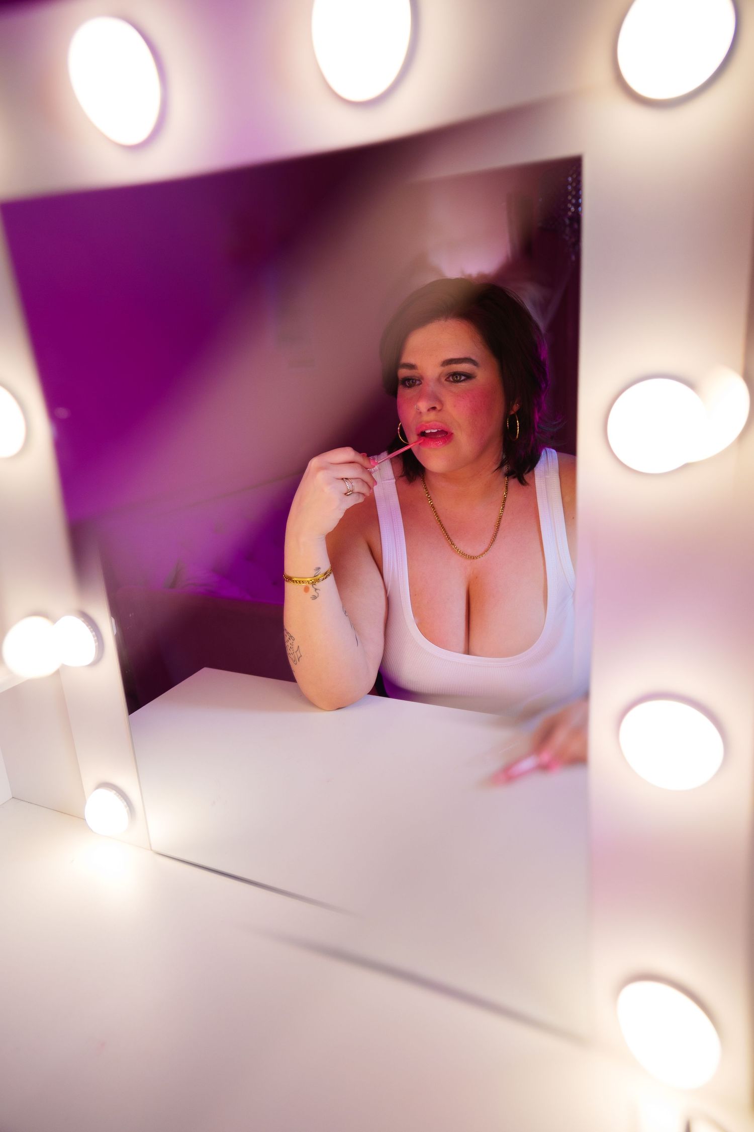 Woman in white tank top applies makeup at a vanity mirror surrounded by Hollywood lights.