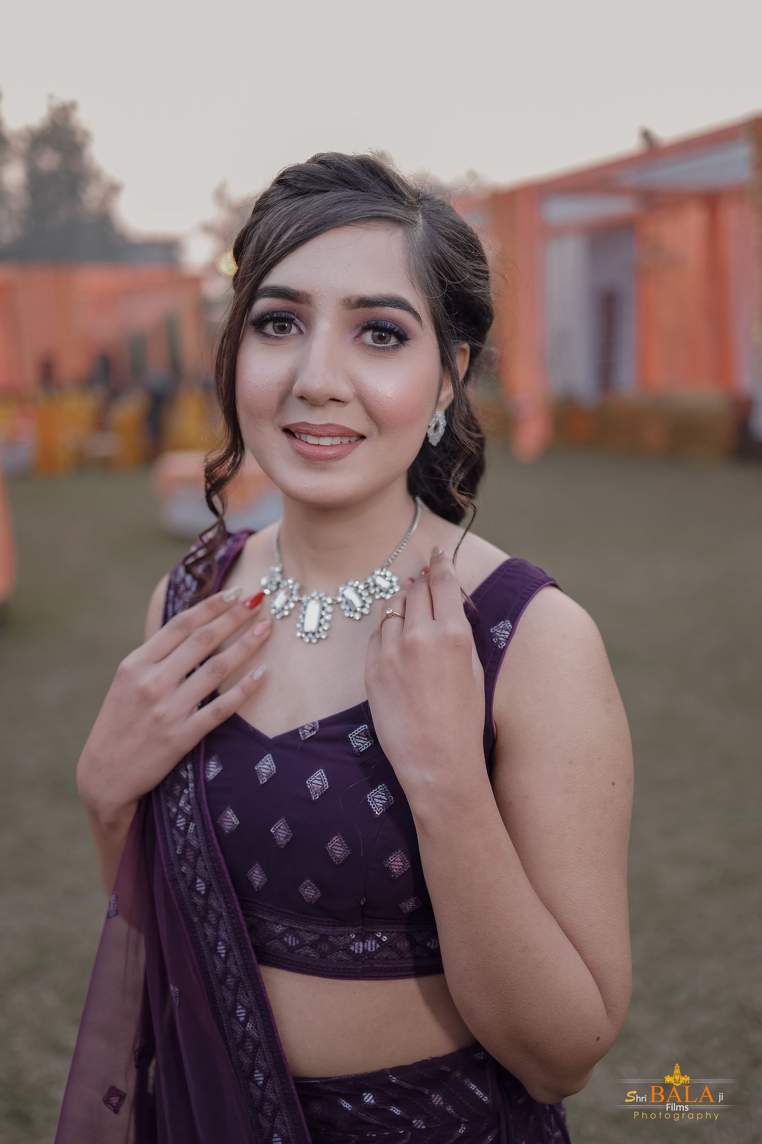 Woman in purple traditional Indian attire with elegant jewelry poses in outdoor setting.