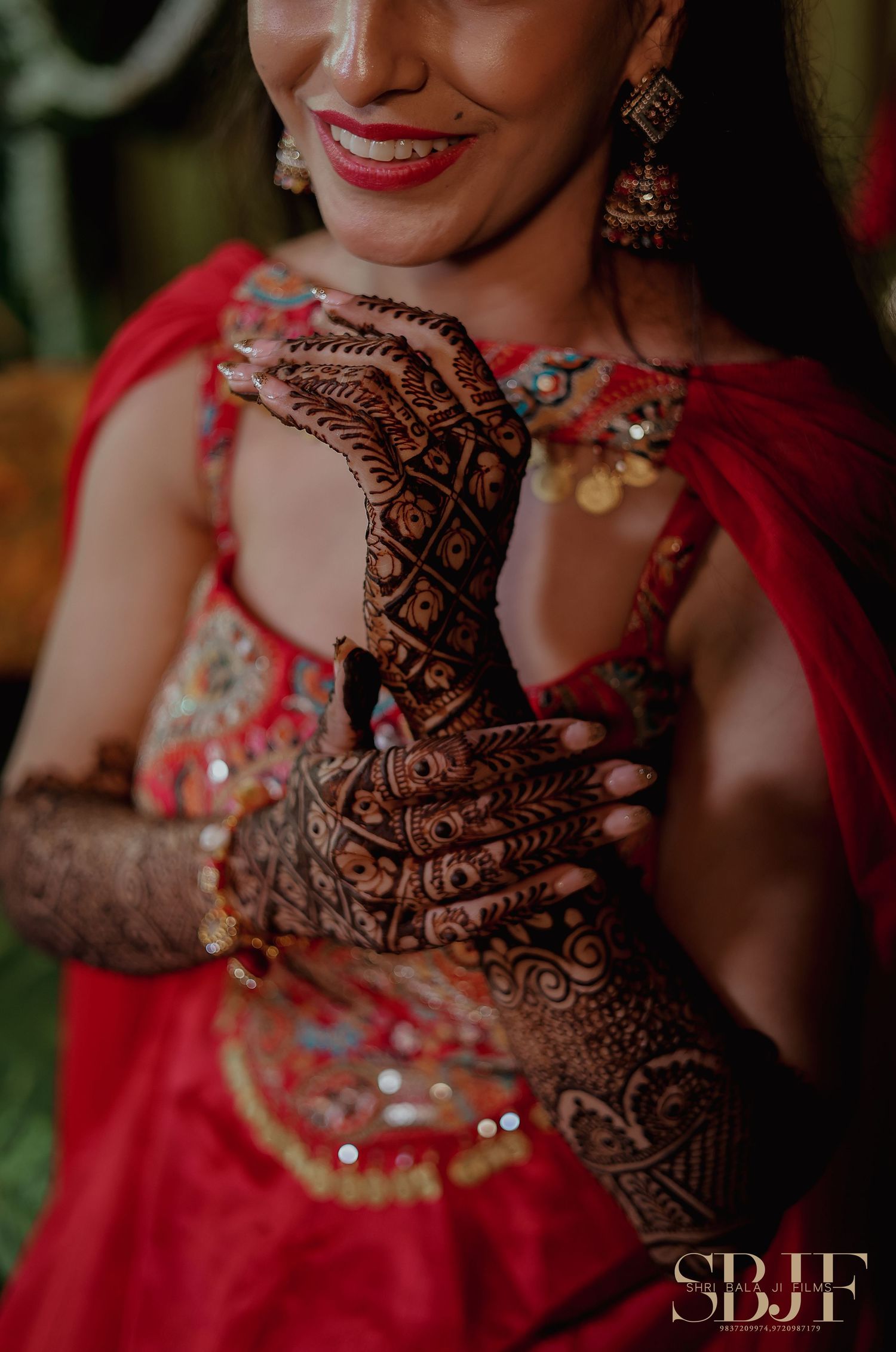 A bride in red traditional attire shows her intricate henna design on her hands with a bright smile and dangling earrings.