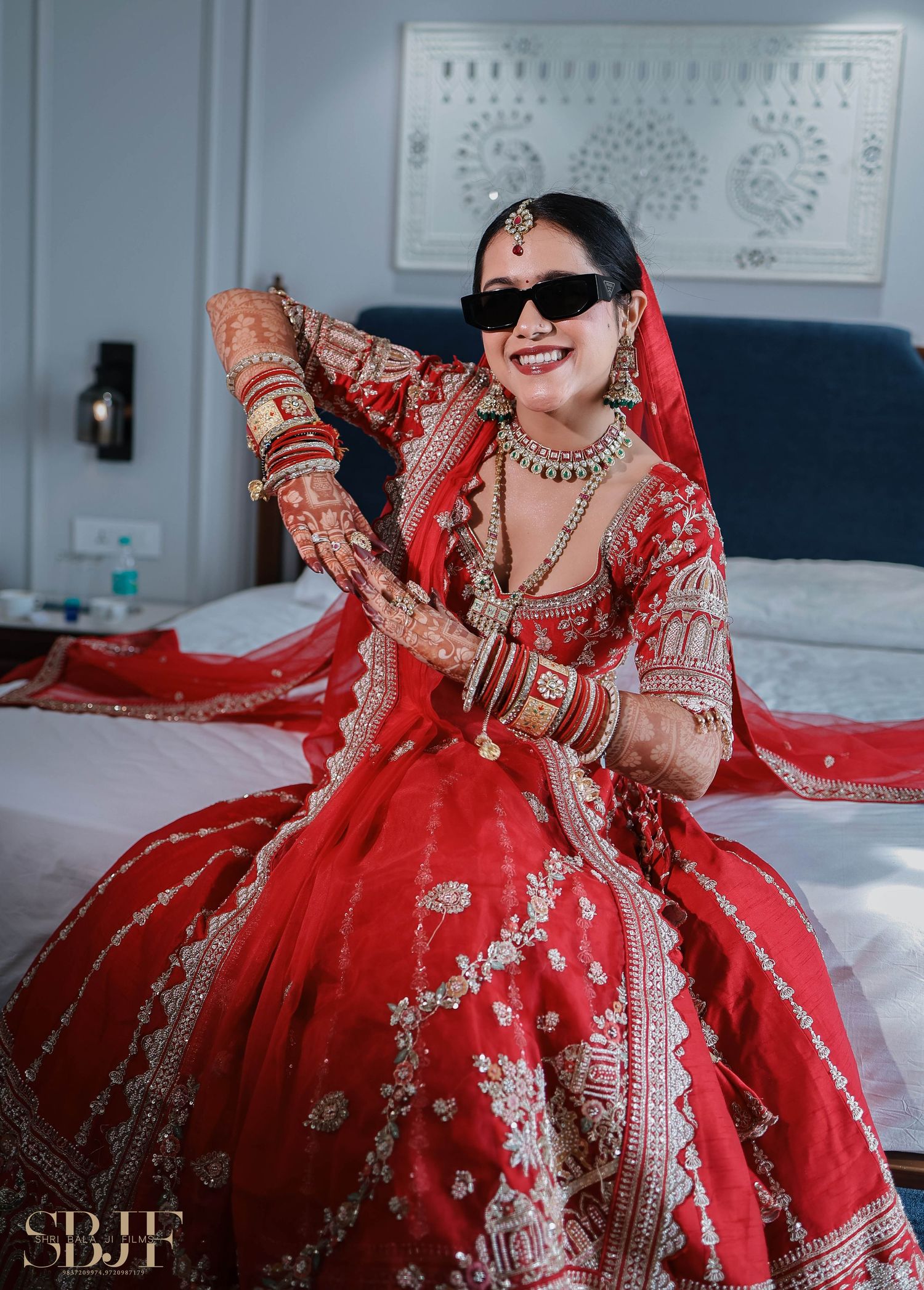 Traditional Indian bride in a vibrant red lehenga with intricate embroidery and sunglasses poses on a bed.
