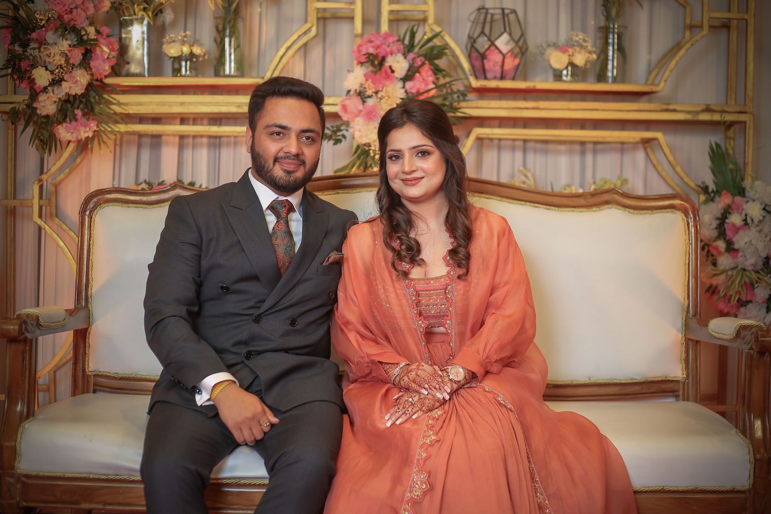 Couple sitting on an ornate couch decorated with pink floral arrangements in an elegant setting.