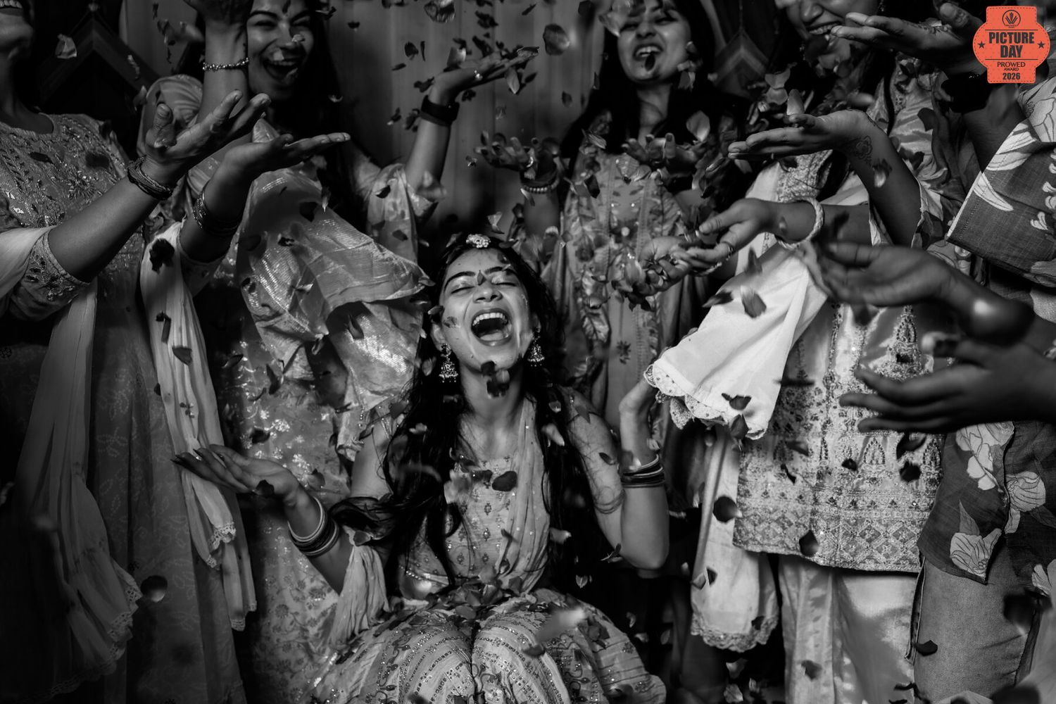 Black and white photo of a joyful bride laughing during a flower shower ceremony surrounded by celebrating guests.