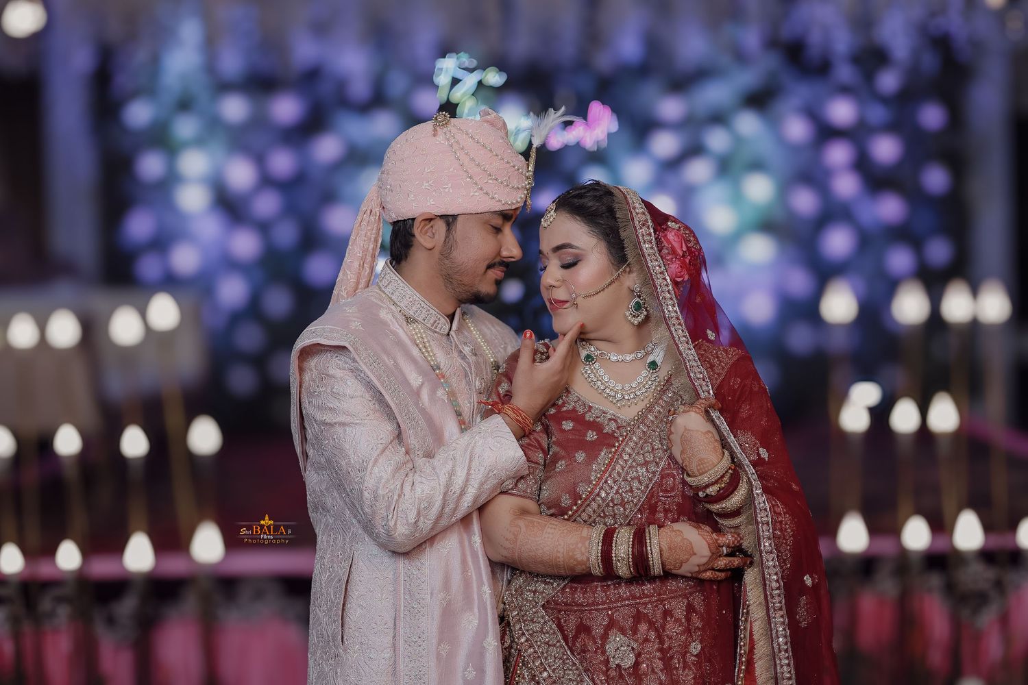 Traditional Indian wedding couple in red and cream outfits against a purple lit backdrop.