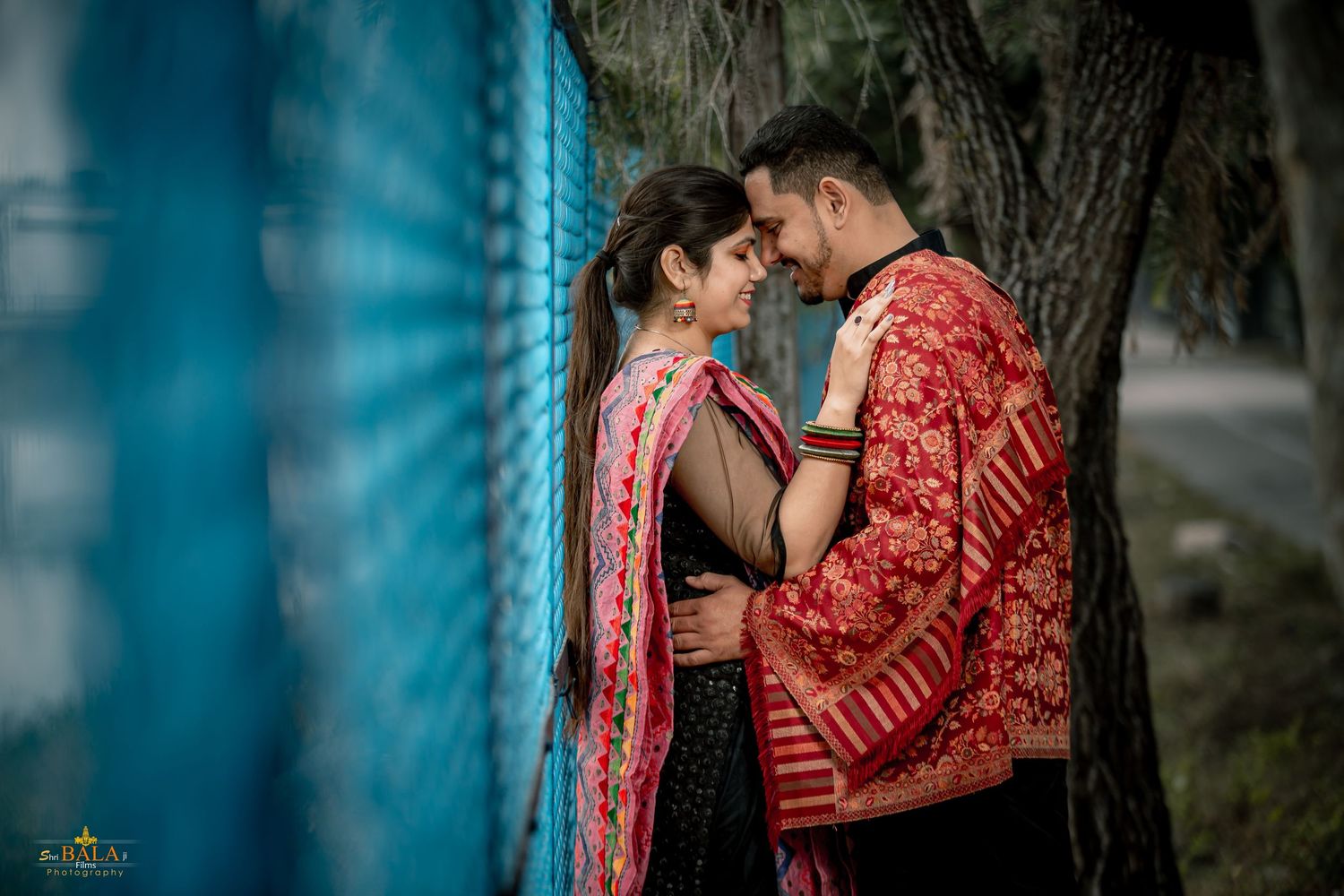 A couple in traditional red wedding attire shares an intimate romantic moment during their outdoor photo session.