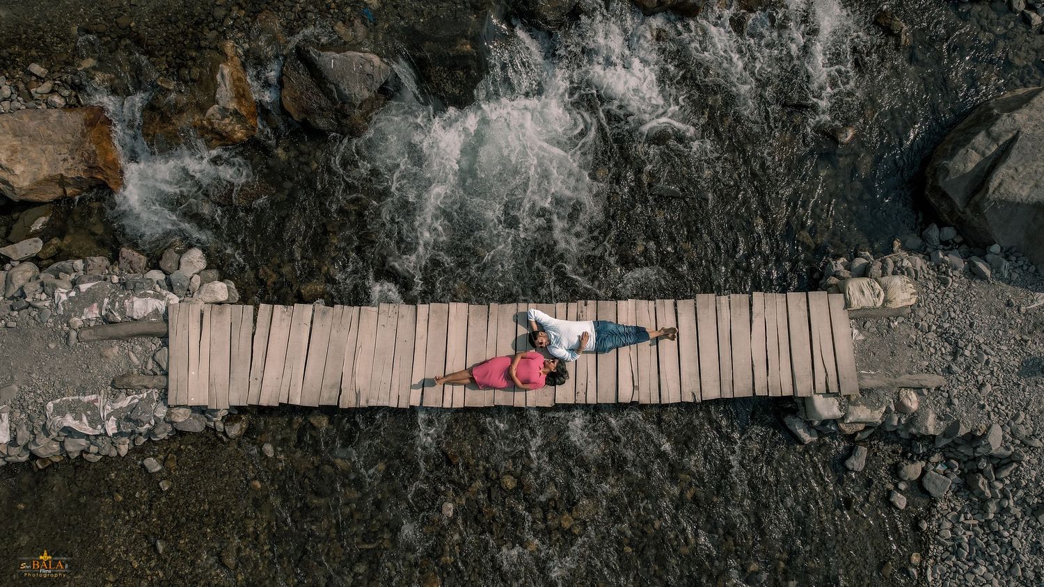 Aerial view of a couple lying on a wooden footbridge surrounded by rocky terrain.