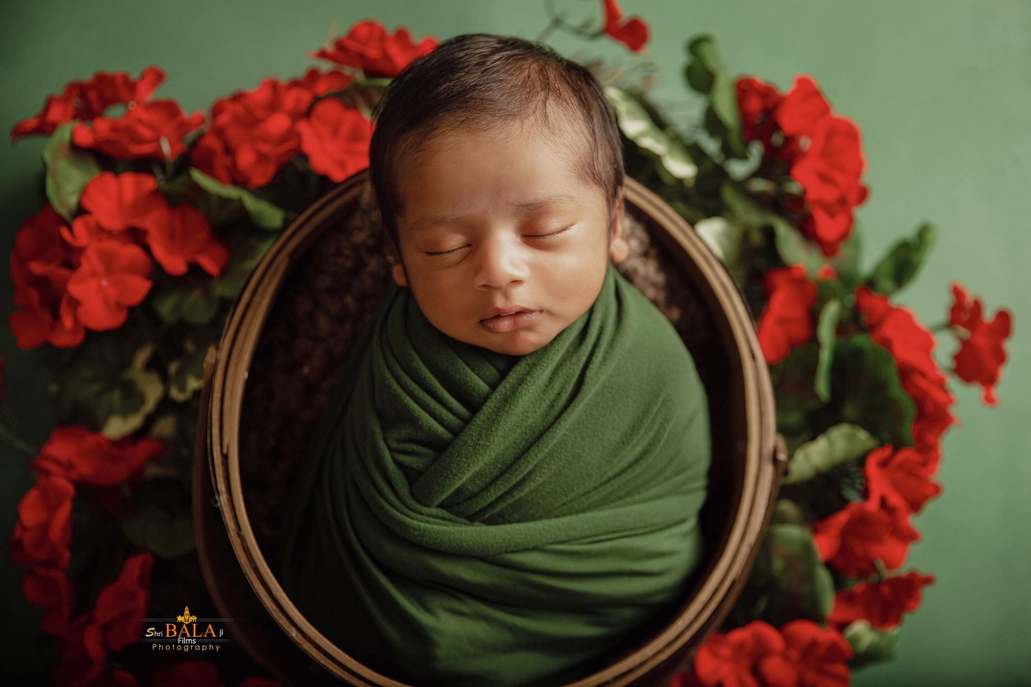 Newborn baby wrapped in dark green fabric nestled in wooden bowl surrounded by vibrant red flowers.
