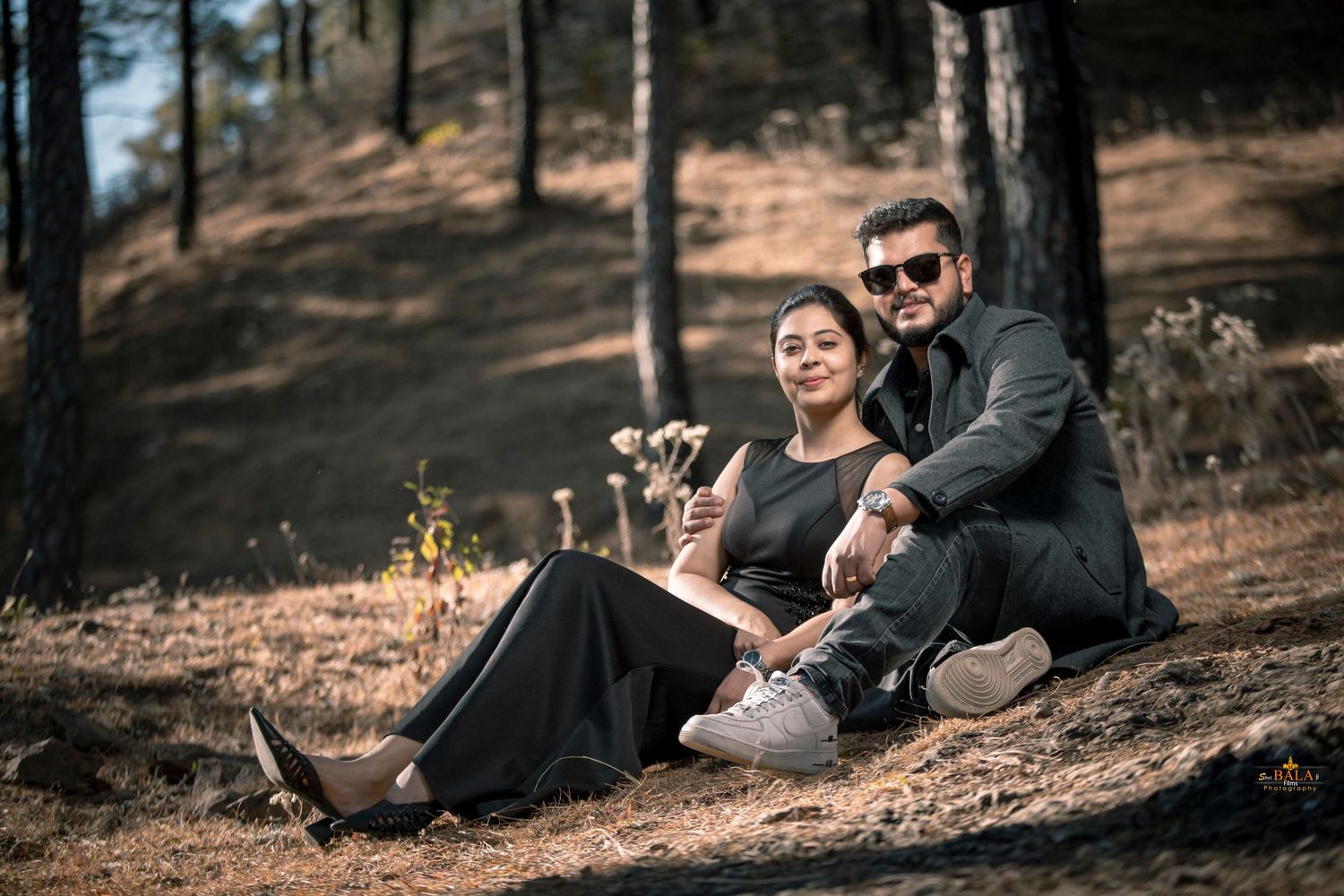 Couple in casual attire relaxing on forest ground during golden hour photoshoot.