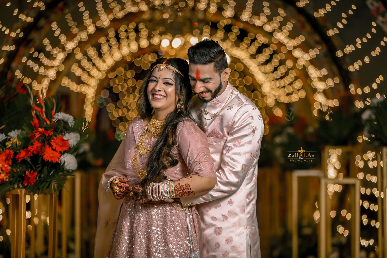 A couple in traditional Indian attire embraces beneath an archway adorned with warm glowing lights and floral decorations.
