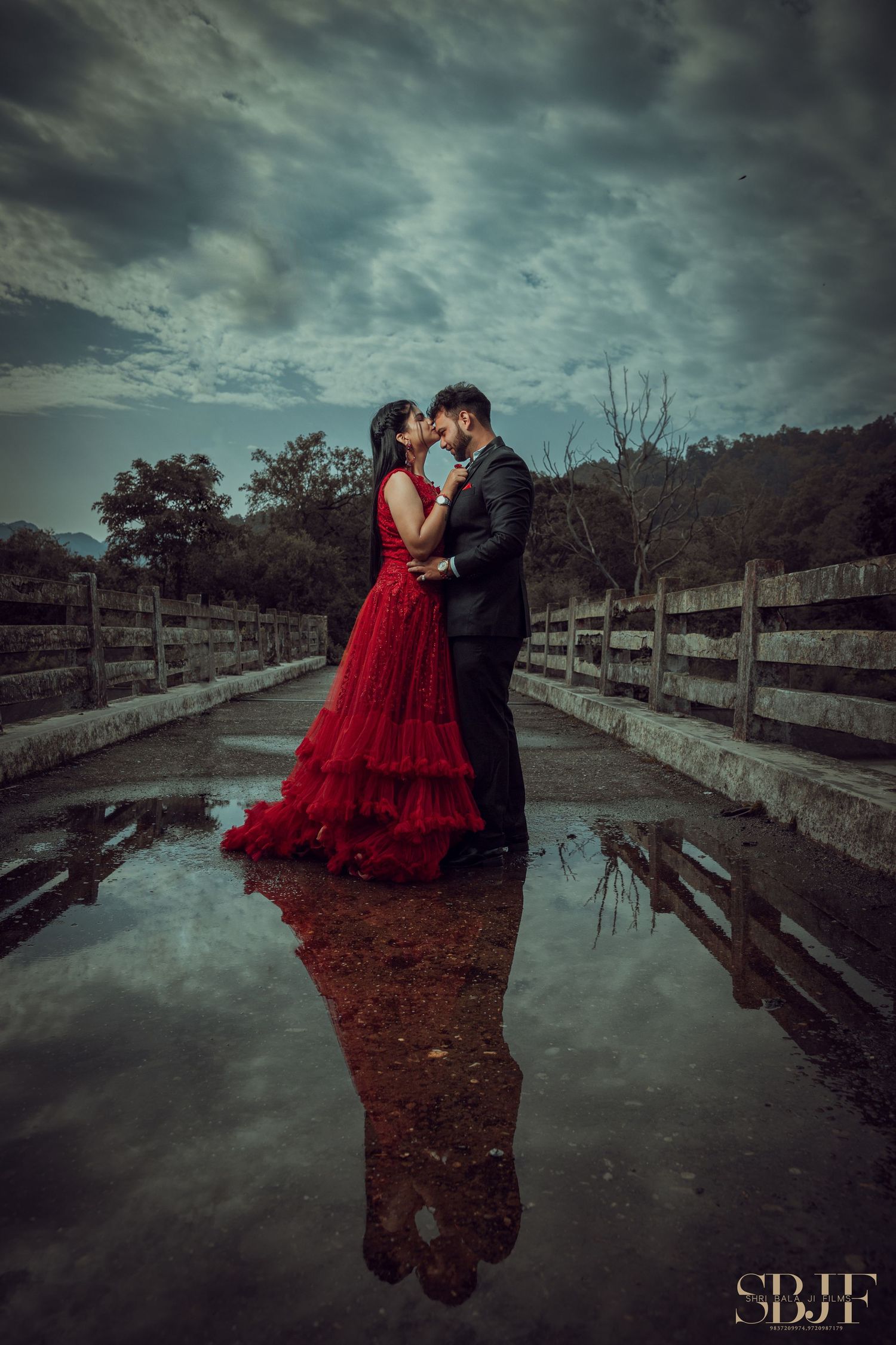 A couple in formal attire shares a romantic moment on a wooden bridge under moody storm clouds at dusk.