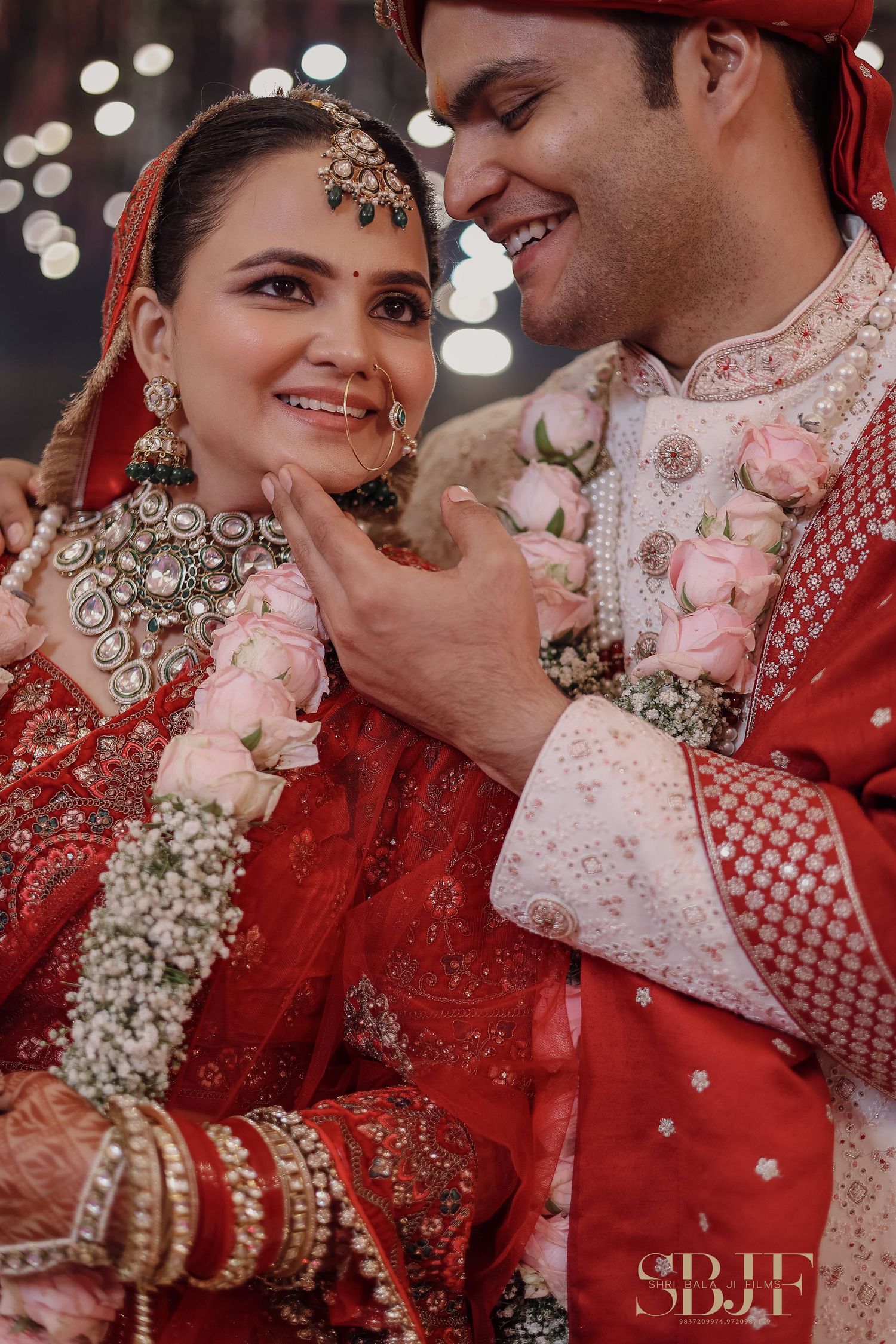 Couple in traditional Indian wedding attire share candid moments during celebration with sparkling lights in background.