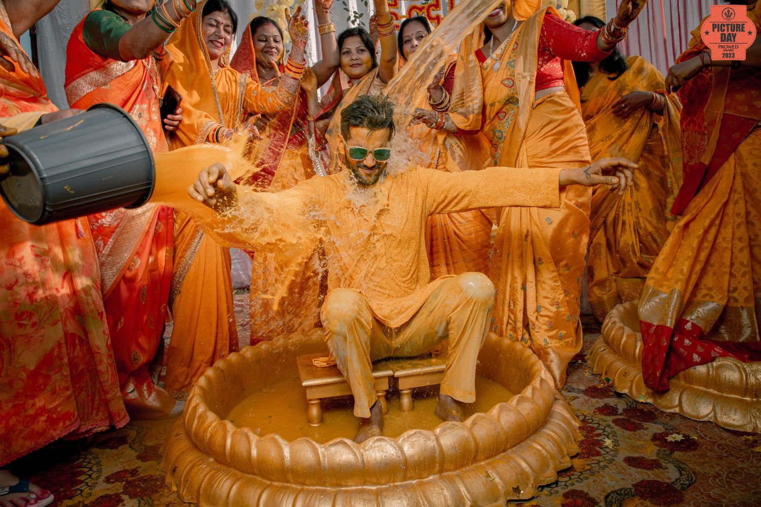 Groom covered in turmeric paste during Haldi ceremony, surrounded by women in orange sarees.