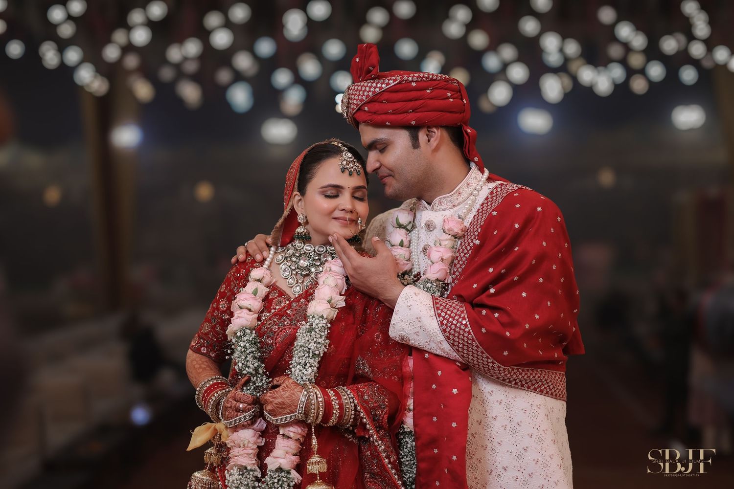 Couple in traditional red wedding attire sharing an intimate moment under glowing string lights.
