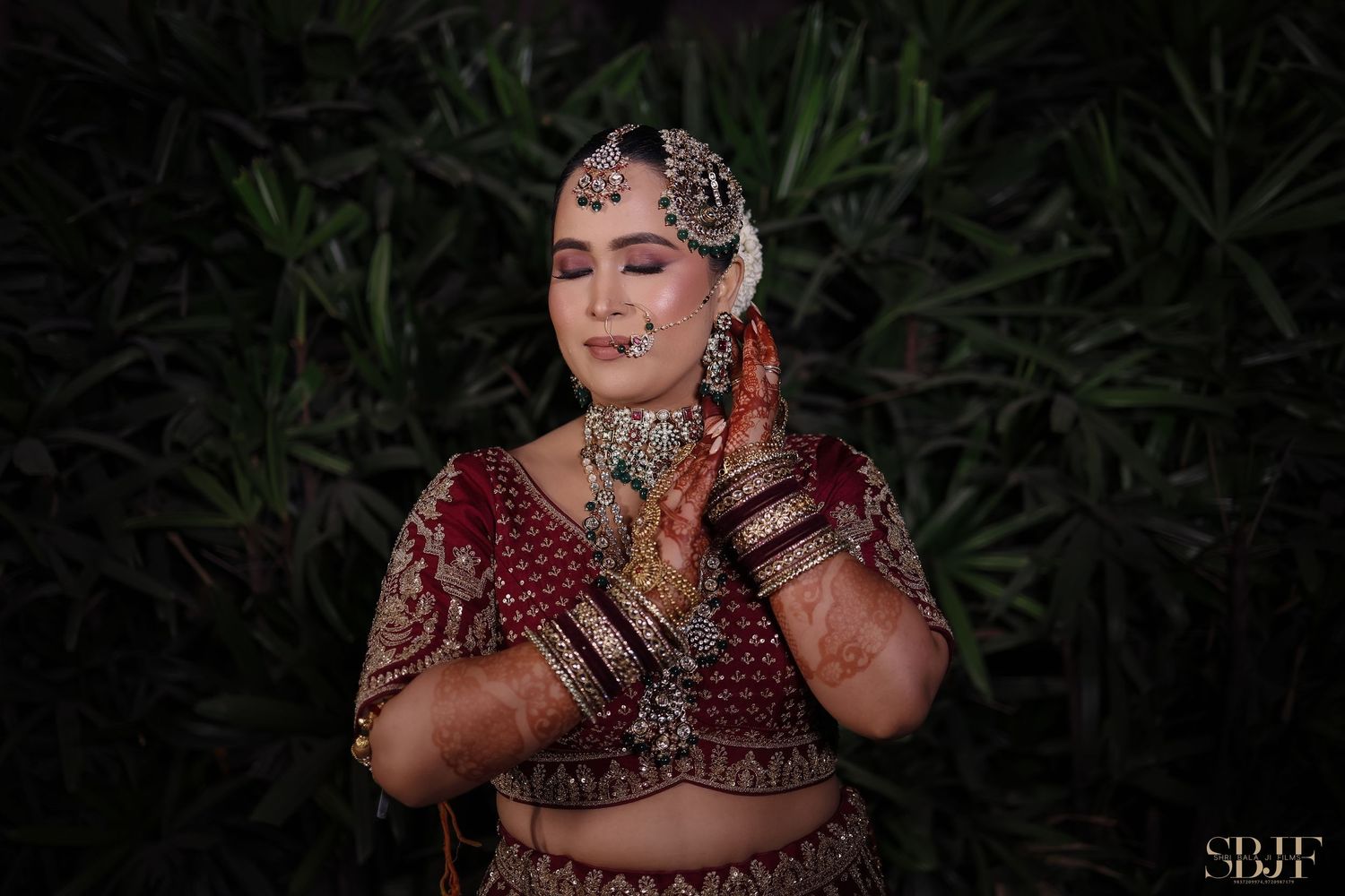 A person in traditional Indian bridal attire with ornate jewelry and henna designs poses against a dark background.