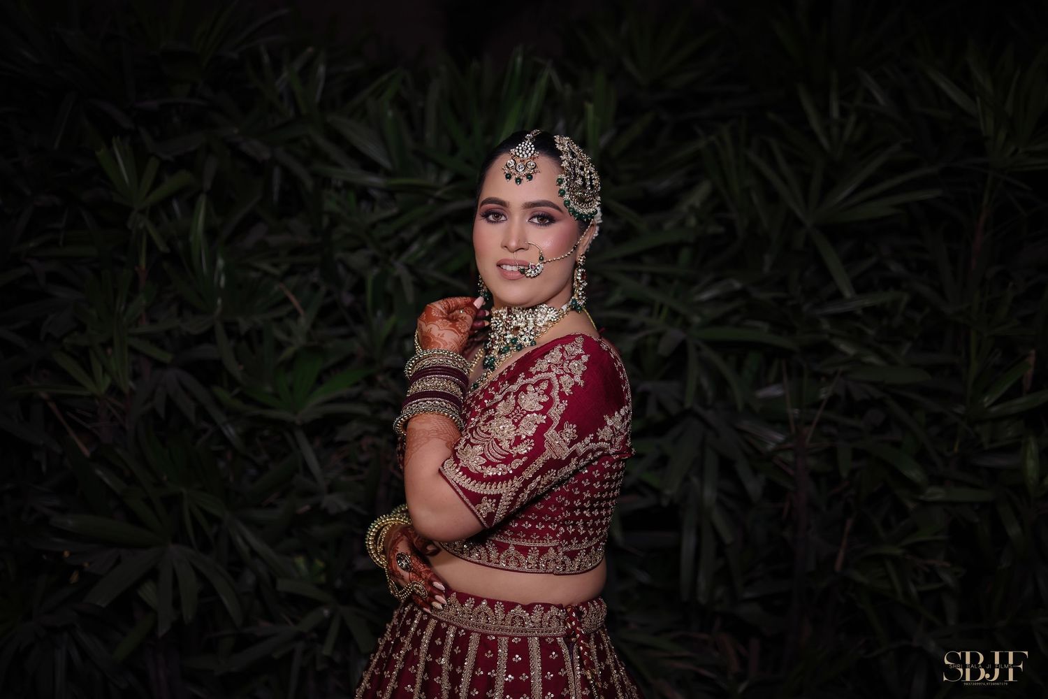A person in an ornate burgundy and gold traditional Indian wedding dress poses in front of dark foliage at night.