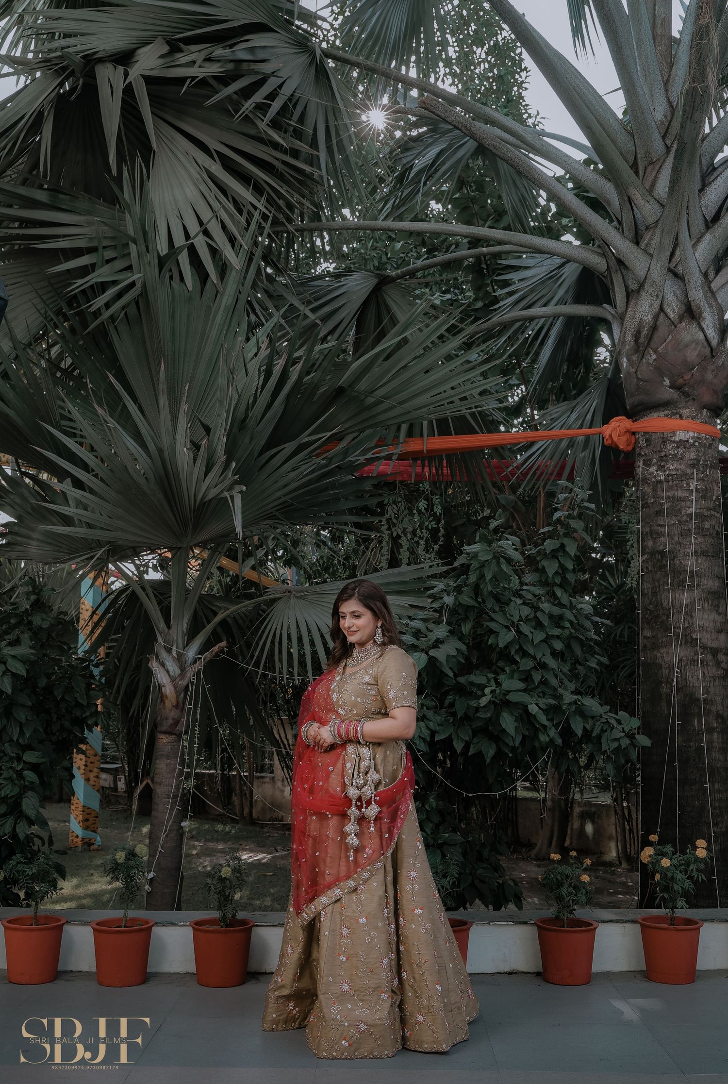 A figure in an ornate gold and red traditional lehenga poses elegantly among tropical palm trees in a garden setting.