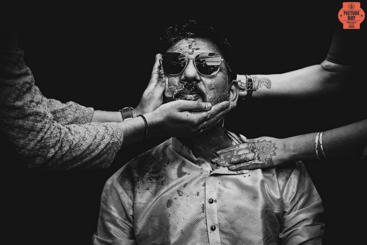 Black and white photo of a man in sunglasses having his face held by henna-adorned hands at a wedding.