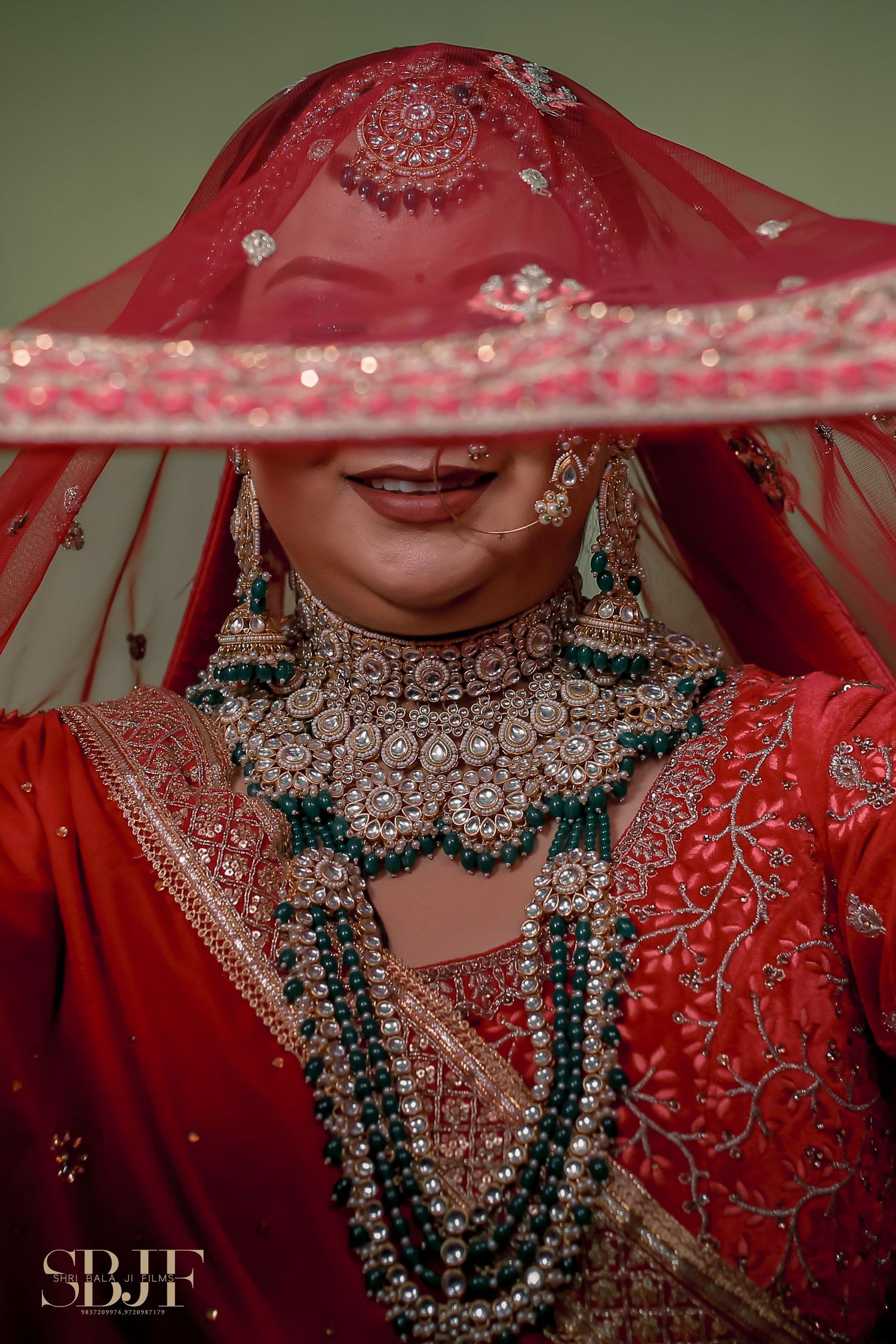 Traditional Indian bride wearing red veil, embellished jewelry and emerald green necklace in artistic portrait.