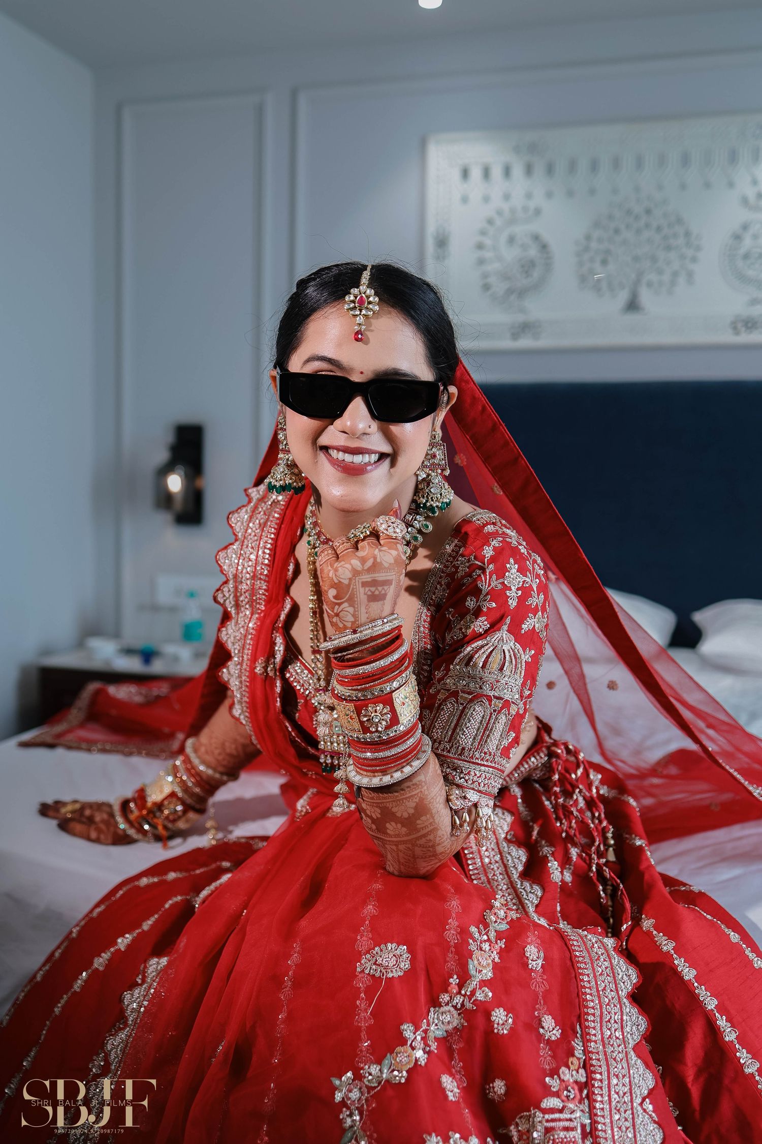 Traditional Indian bride wearing a vibrant red lehenga with ornate gold embroidery and jewelry poses on a bed.