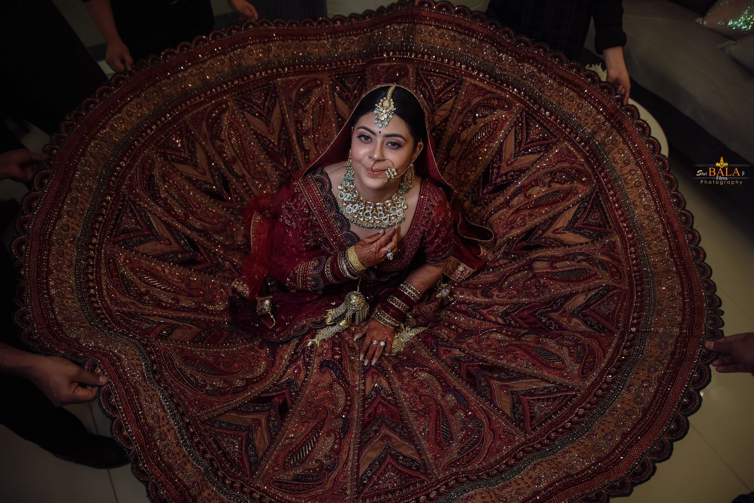 A bride in a deep red traditional Indian lehenga with ornate jewelry sits on an intricately patterned floor.