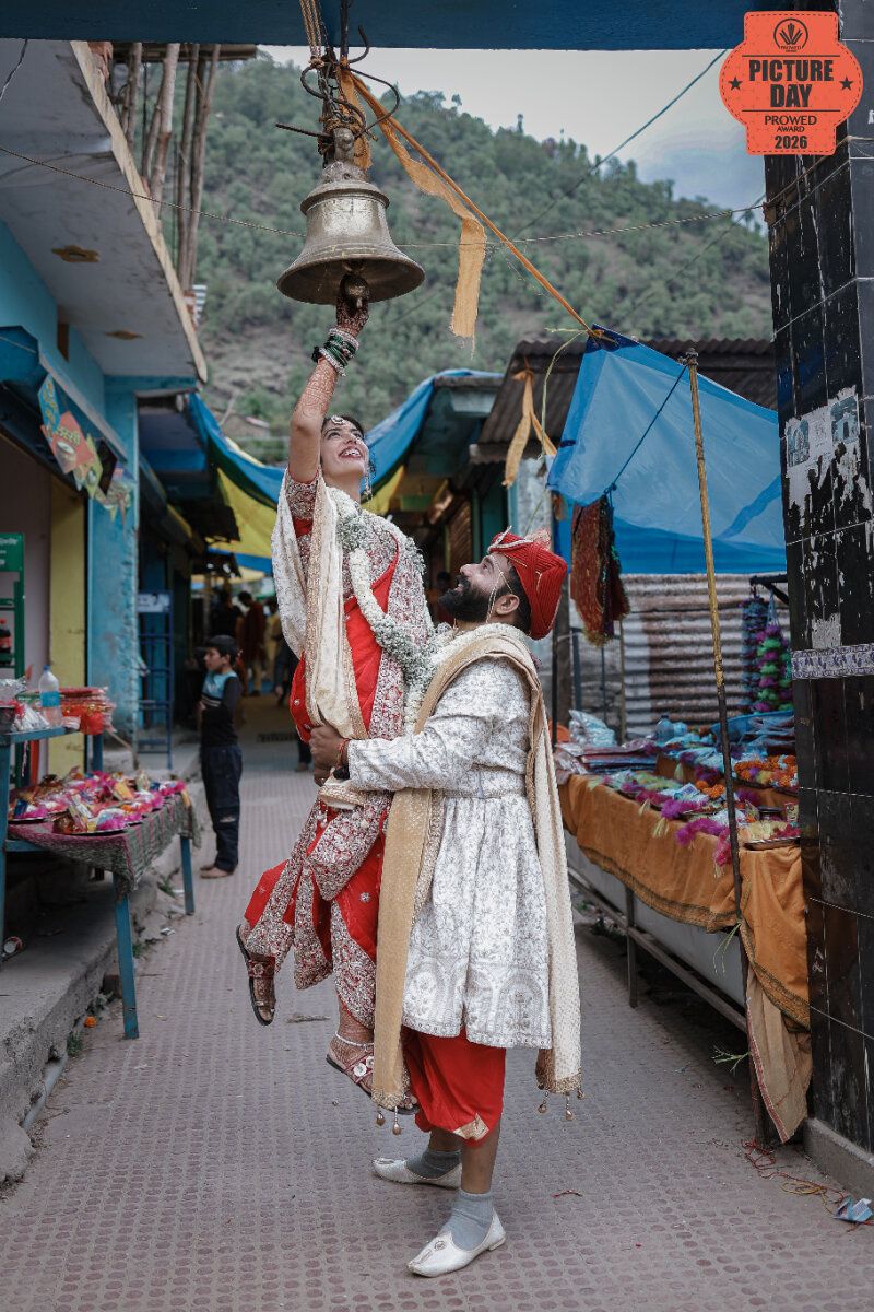 Indian bride and groom in traditional attire ringing a temple bell in a colorful mountain market street.