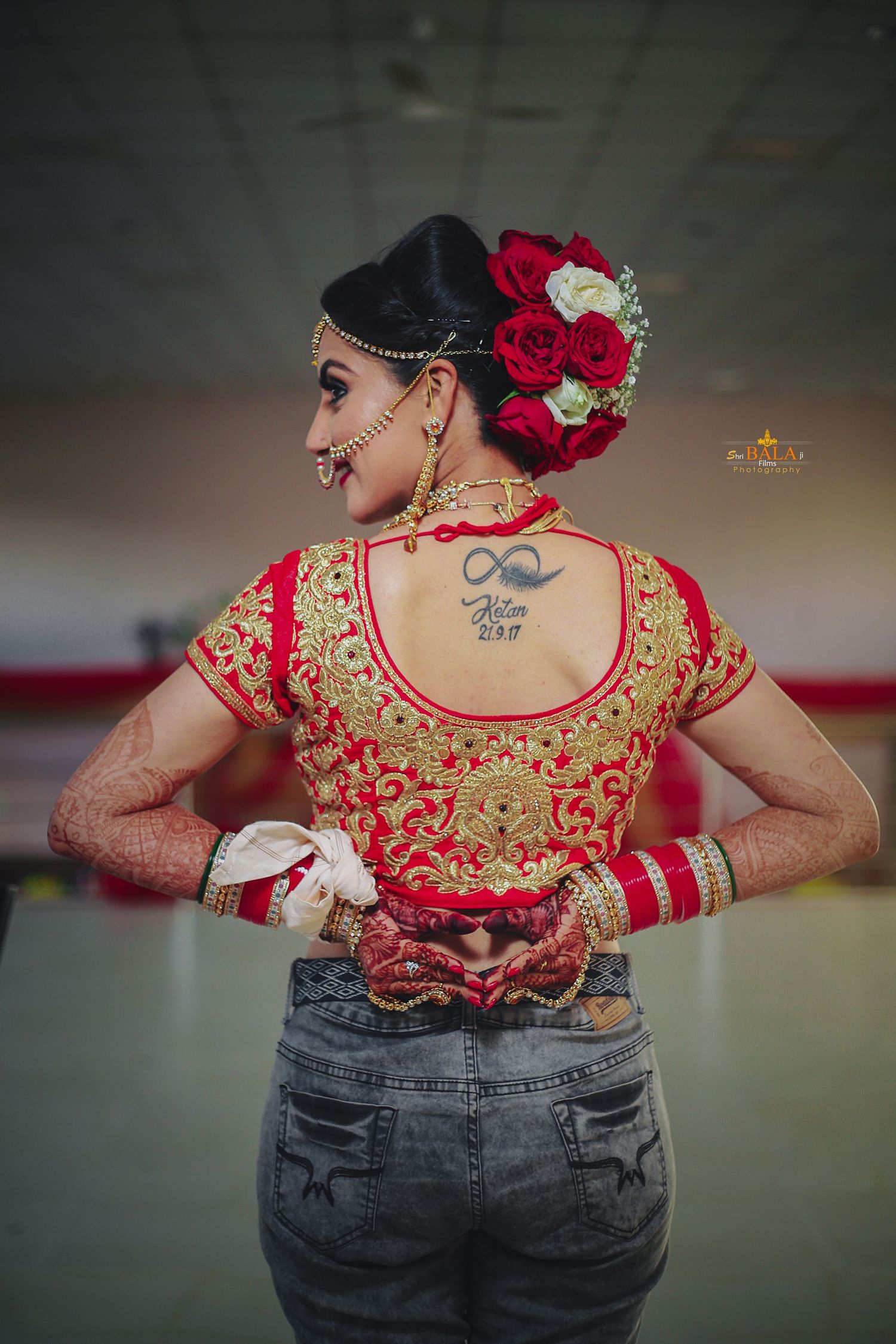 Back view of bride wearing red and gold embroidered blouse with roses in hair and showing traditional mehndi on hands.