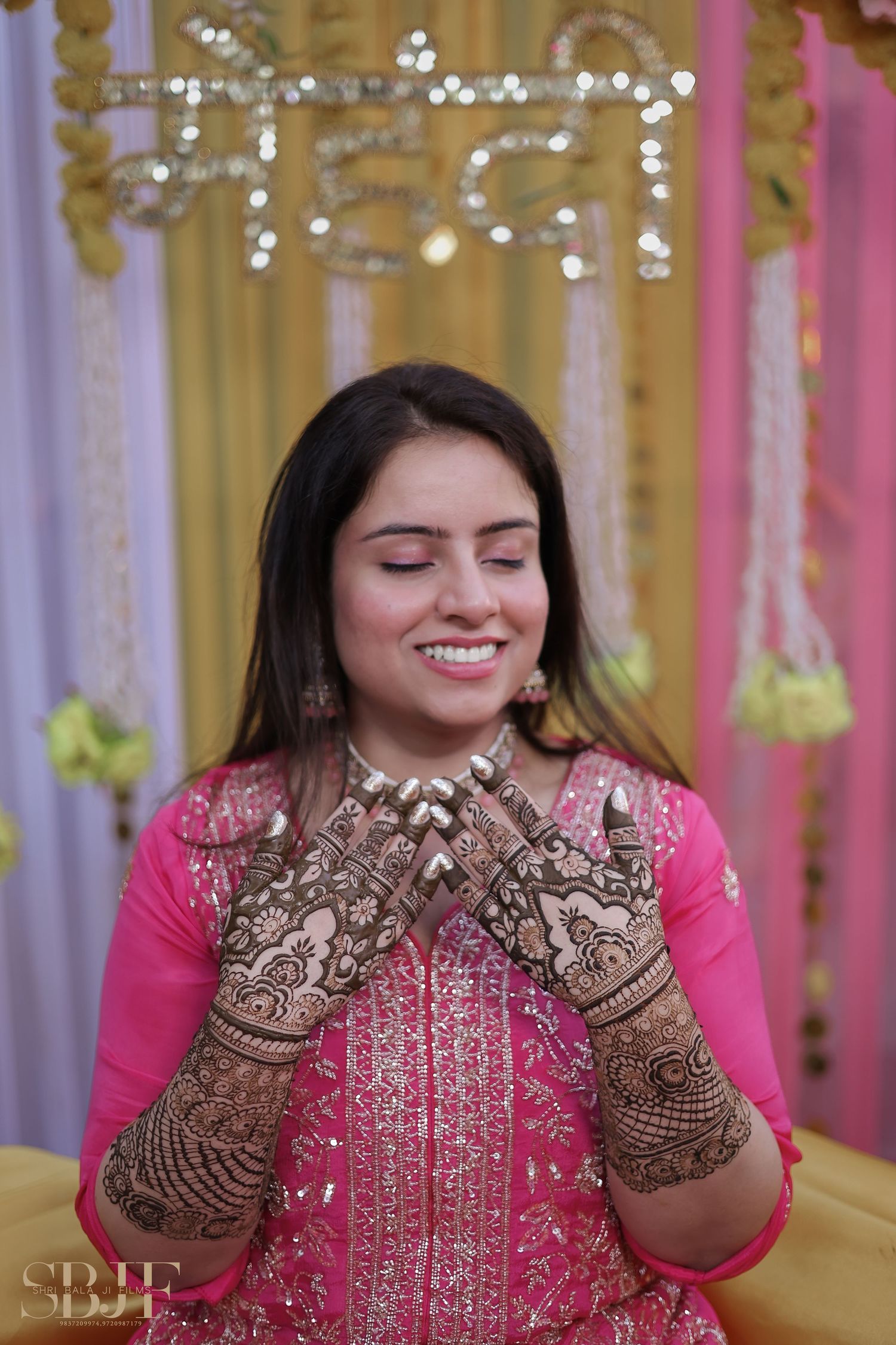 Wedding guest wearing pink traditional outfit shows off intricate henna designs on hands with decorative backdrop.