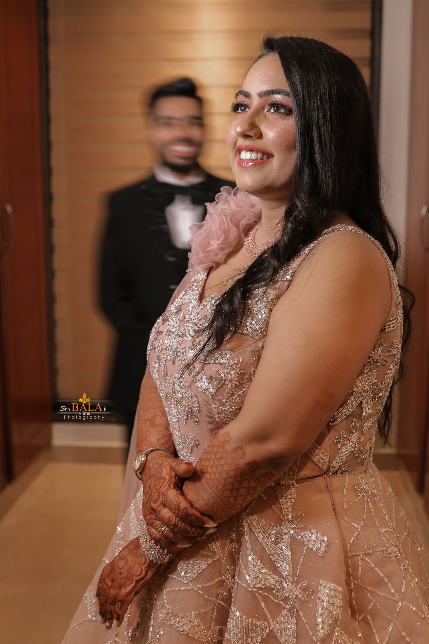A woman in a blush pink embellished gown smiles while showing her henna-adorned hands at a formal event.