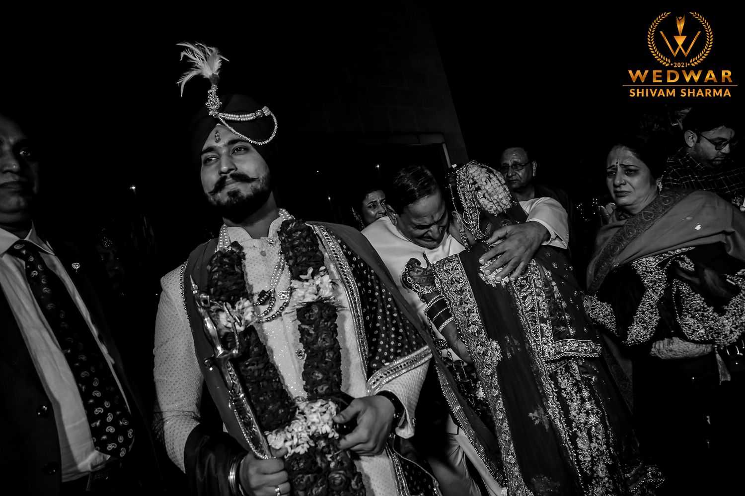 Groom in traditional Sikh wedding attire with floral garlands walks with guests at Indian wedding ceremony.